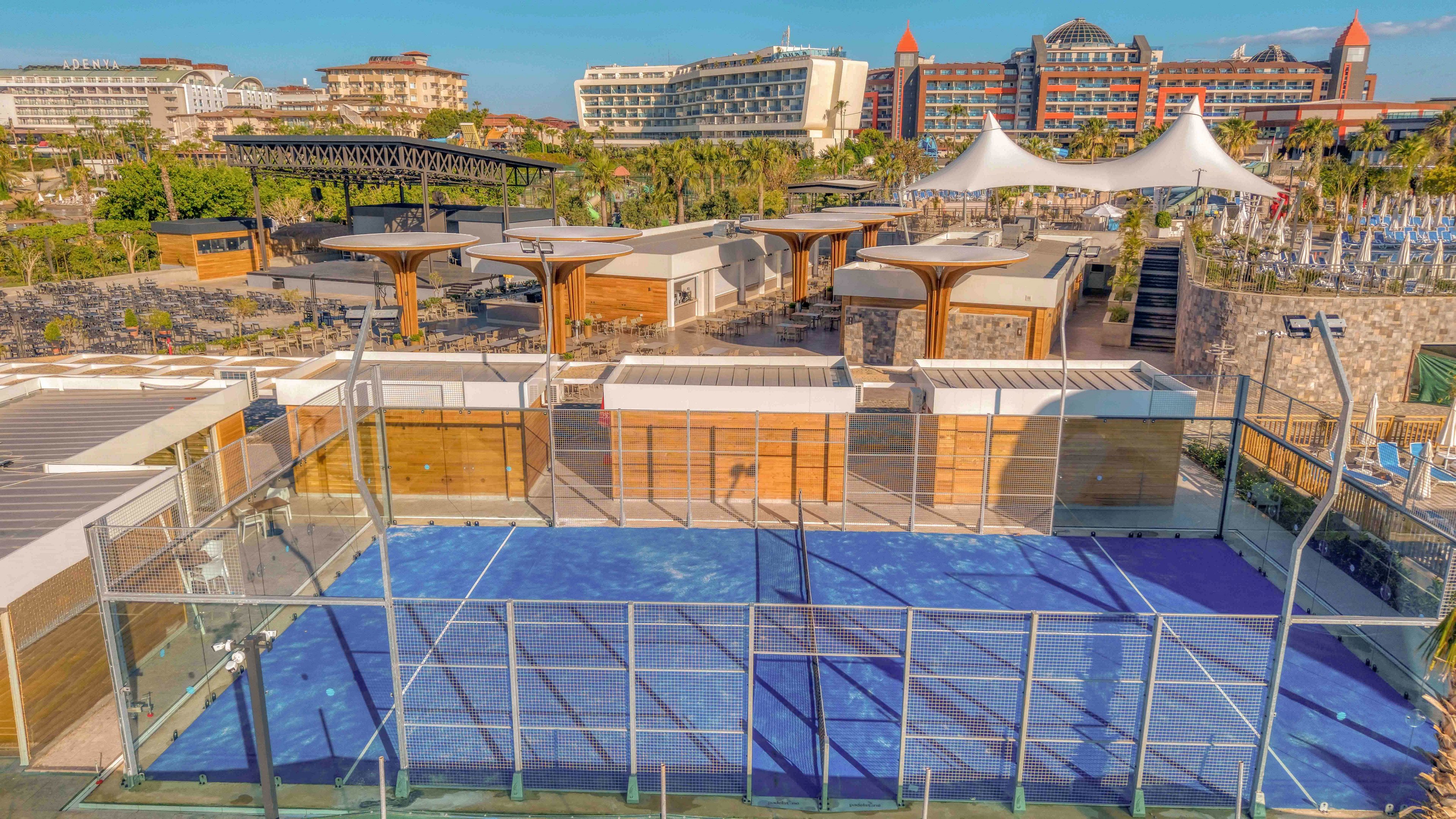 An elevated view shows a blue outdoor padel court enclosed by metal fencing in a resort setting. Surrounding the court are wooden cabanas, shaded seating areas, and landscaped walkways. In the background, large hotel buildings and white tensile canopies overlook the recreational area. The scene is brightly lit with clear skies, emphasizing a sunny vacation atmosphere.