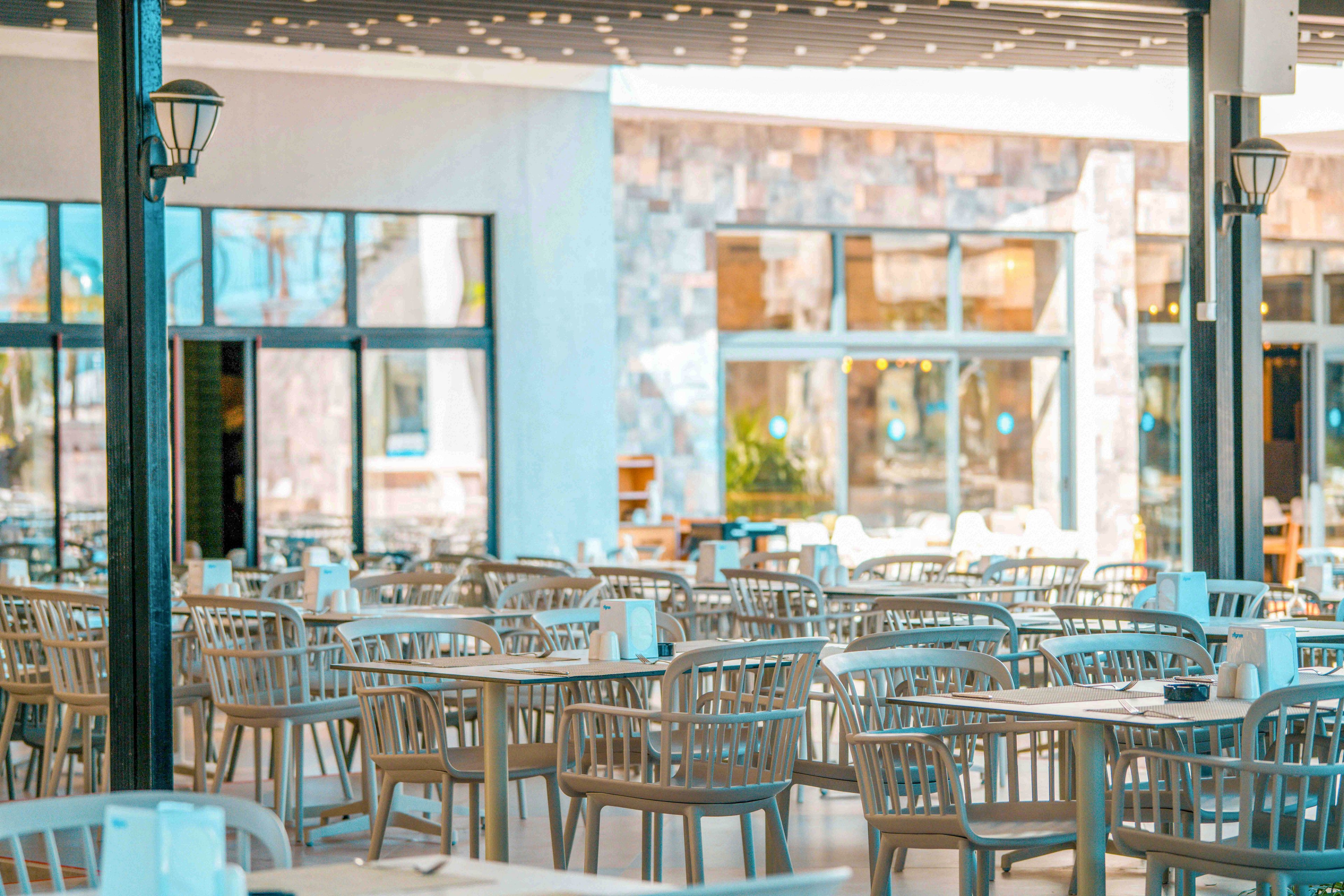An open-air restaurant terrace is filled with rows of empty tables and matching chairs. The setting appears bright and sunny, with warm light reflecting off the stone wall and large glass windows in the background. Tabletop items such as napkin dispensers and condiments are neatly arranged, suggesting the space is prepared for guests. Overhead lighting and structural columns frame the relaxed, modern dining area.