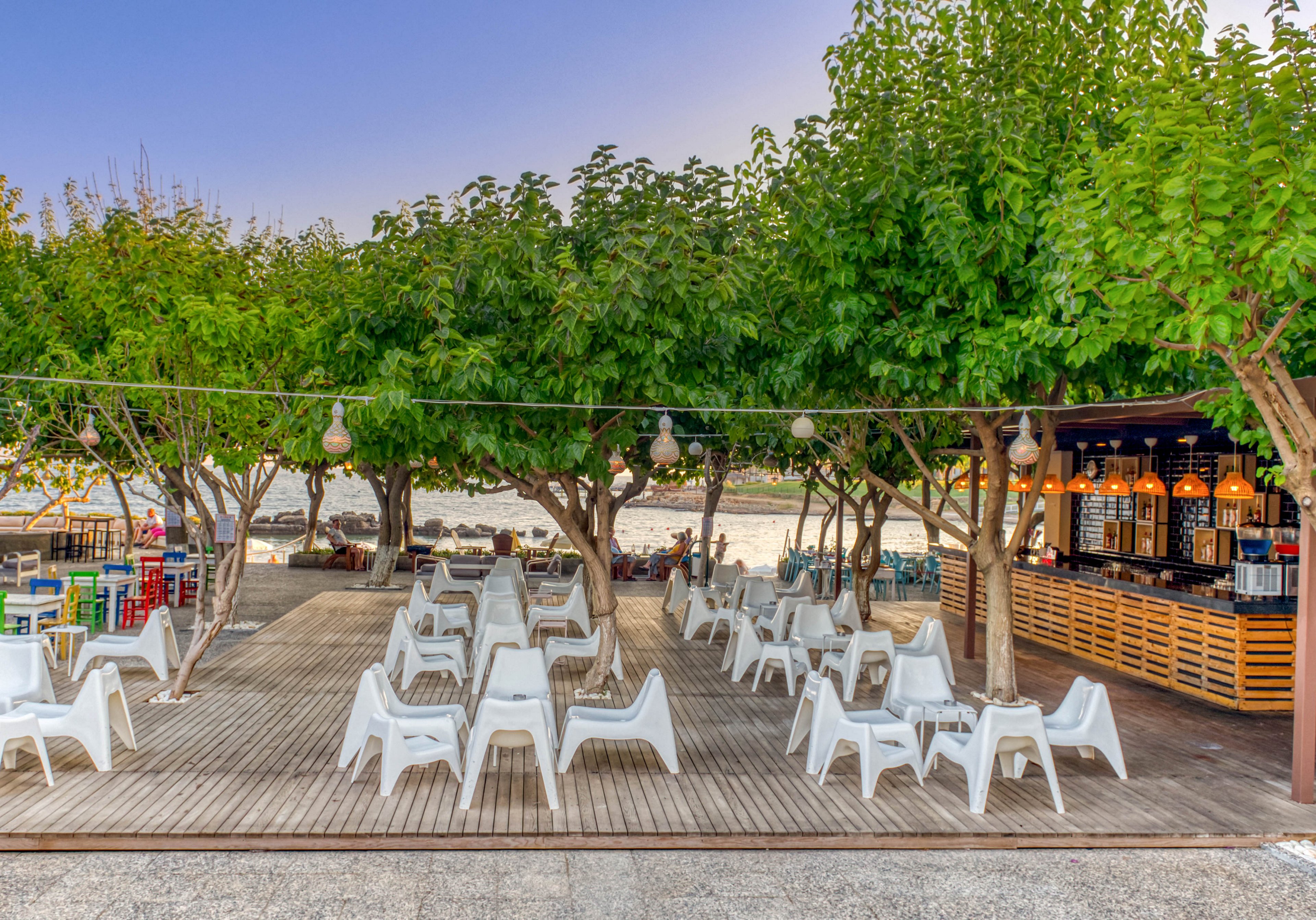 An open-air beach bar terrace features rows of white plastic chairs and tables arranged on a wooden deck. Lush green trees provide shade, with hanging lights strung between them, creating a cozy atmosphere. The bar area on the right side is warmly lit with decorative pendant lamps and stocked shelves. In the background, the sandy beach and calm water are visible, suggesting a relaxed seaside setting.