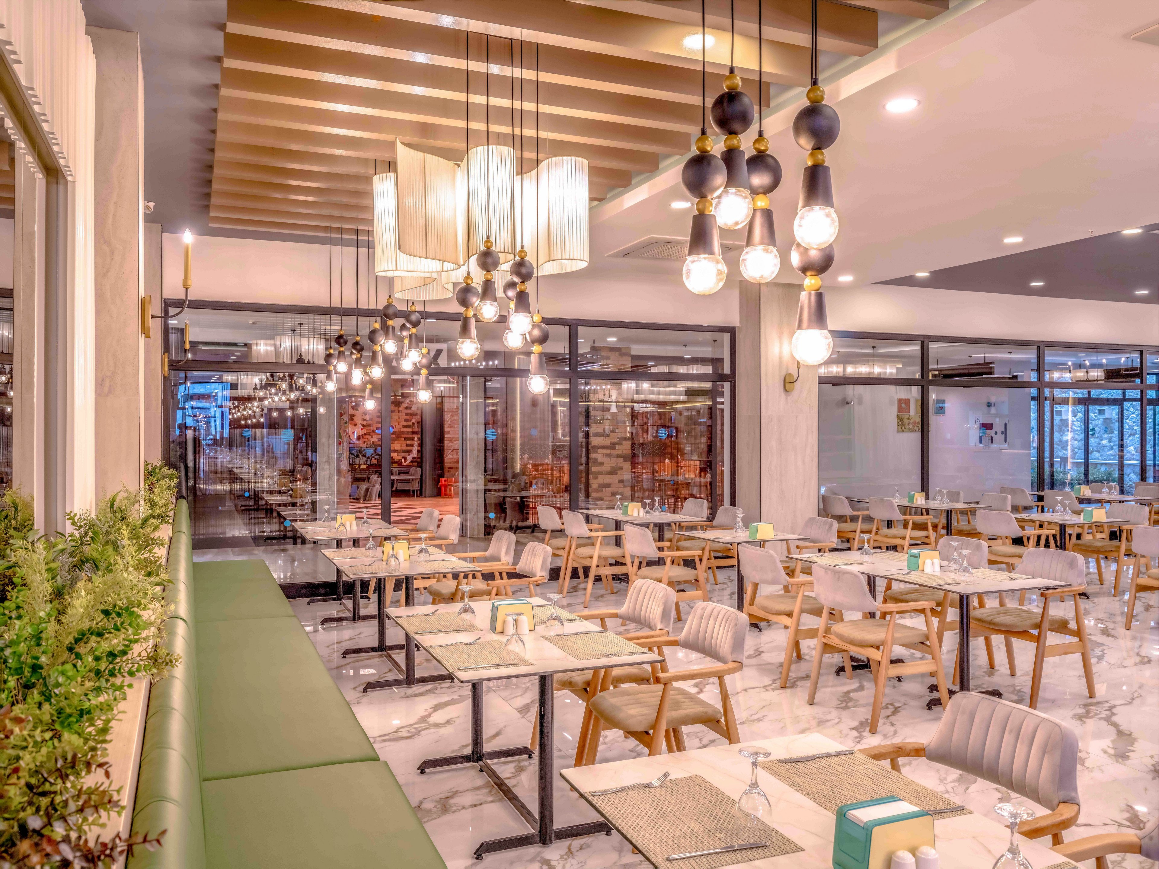 A contemporary restaurant interior features neatly arranged wooden tables and chairs on a glossy marble floor. Multiple decorative pendant lights hang from the ceiling, creating a warm and inviting ambiance. A long green upholstered bench runs along one side, complemented by potted greenery and neutral-toned walls. Large glass windows and doors open to another dining area, enhancing the spacious, airy feel of the space.