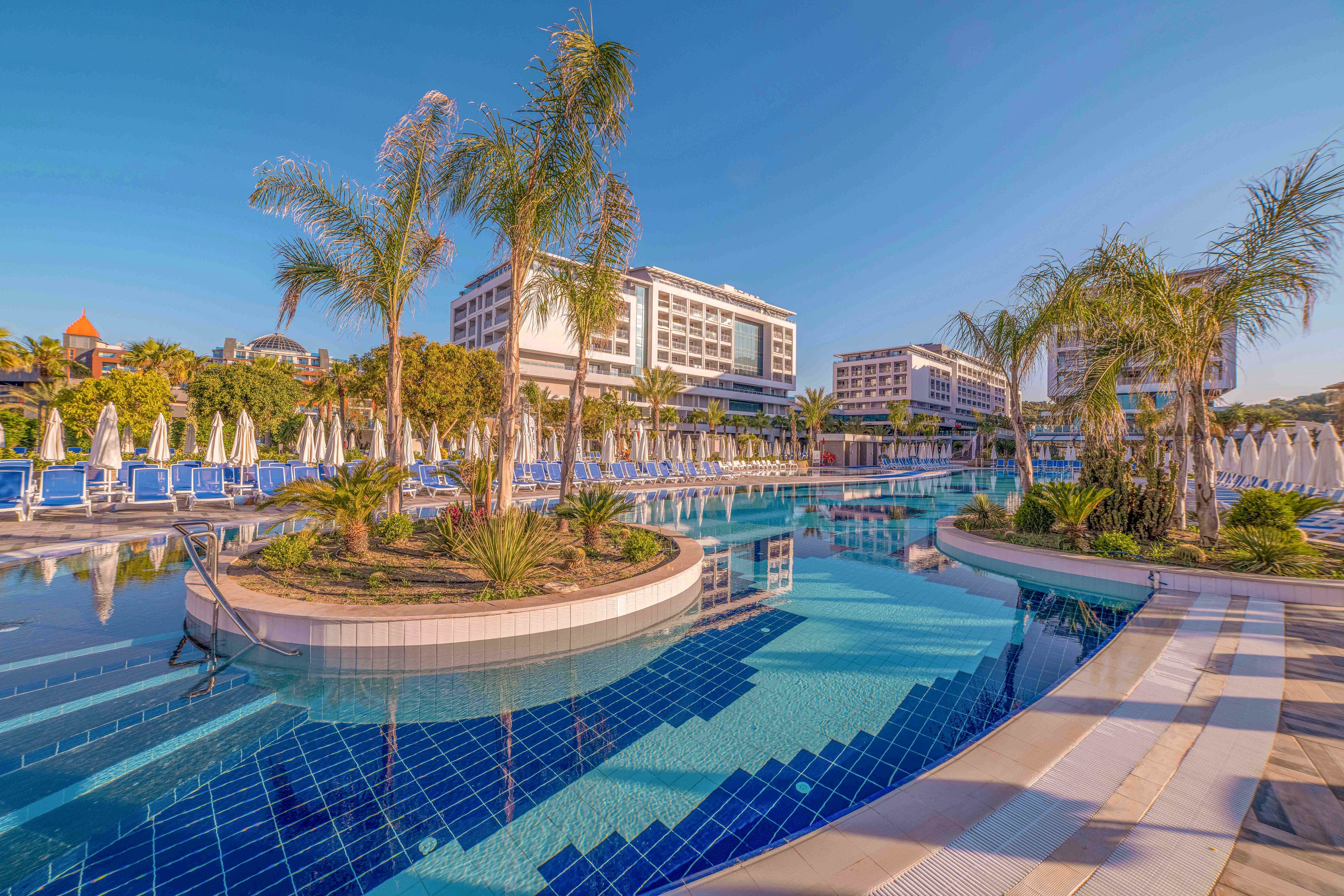 A large resort hotel complex features an expansive outdoor swimming pool surrounded by modern multi-story buildings. Palm trees and landscaped islands decorate the pool area, creating a tropical vacation atmosphere. Numerous sun loungers and closed umbrellas line the poolside, suggesting a relaxing leisure setting. The clear blue sky and reflections in the water emphasize the warm, sunny climate and inviting environment.
