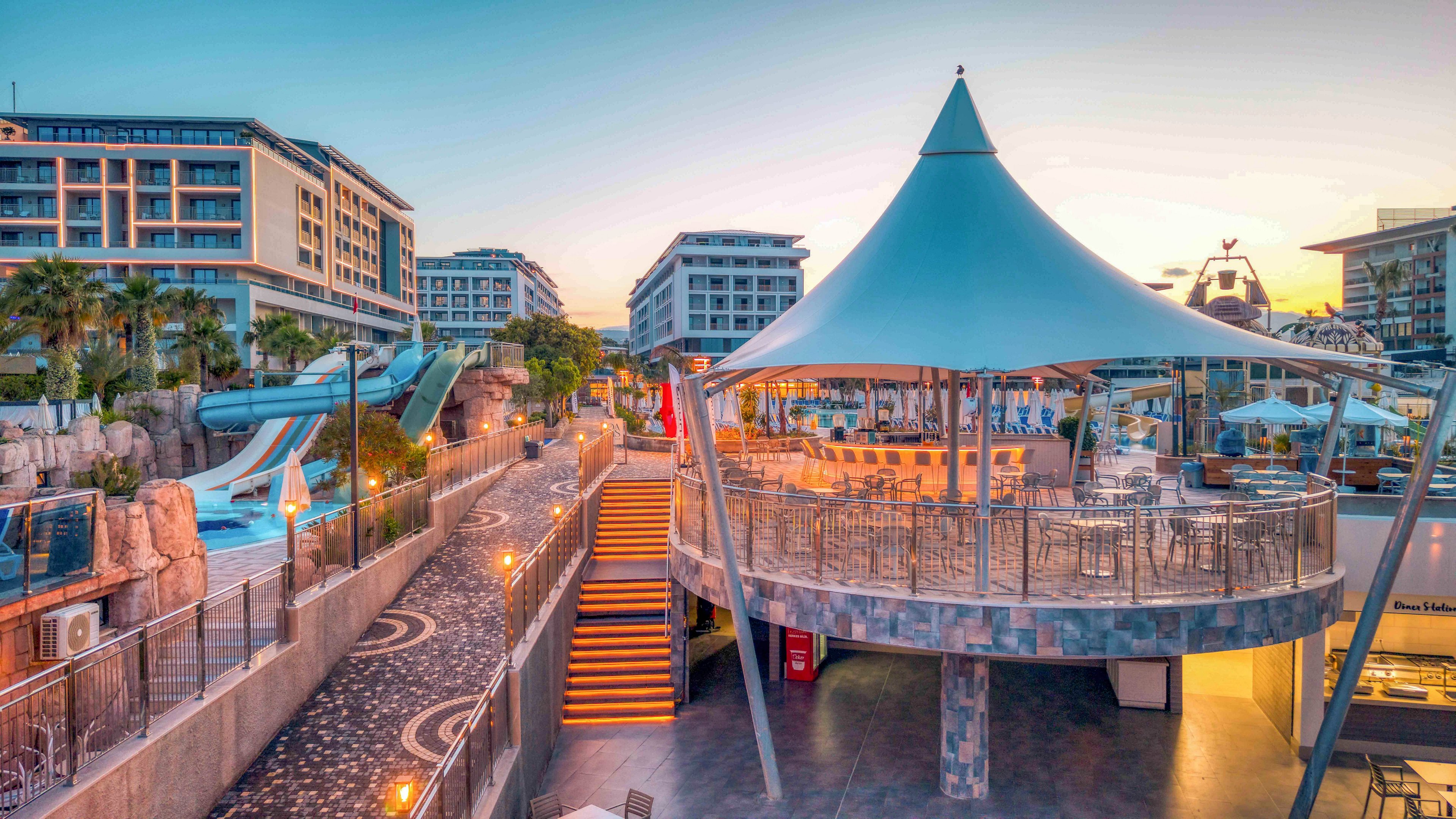 A contemporary seaside resort complex is shown at sunset, featuring a large tented terrace structure in the foreground. Multiple hotel buildings surround an outdoor pool area with tall waterslides and rock-style landscaping. Warm artificial lighting contrasts with the soft blue and orange tones of the evening sky, creating a relaxed vacation atmosphere. The scene is empty of people, emphasizing the architecture, leisure facilities, and tranquil mood.