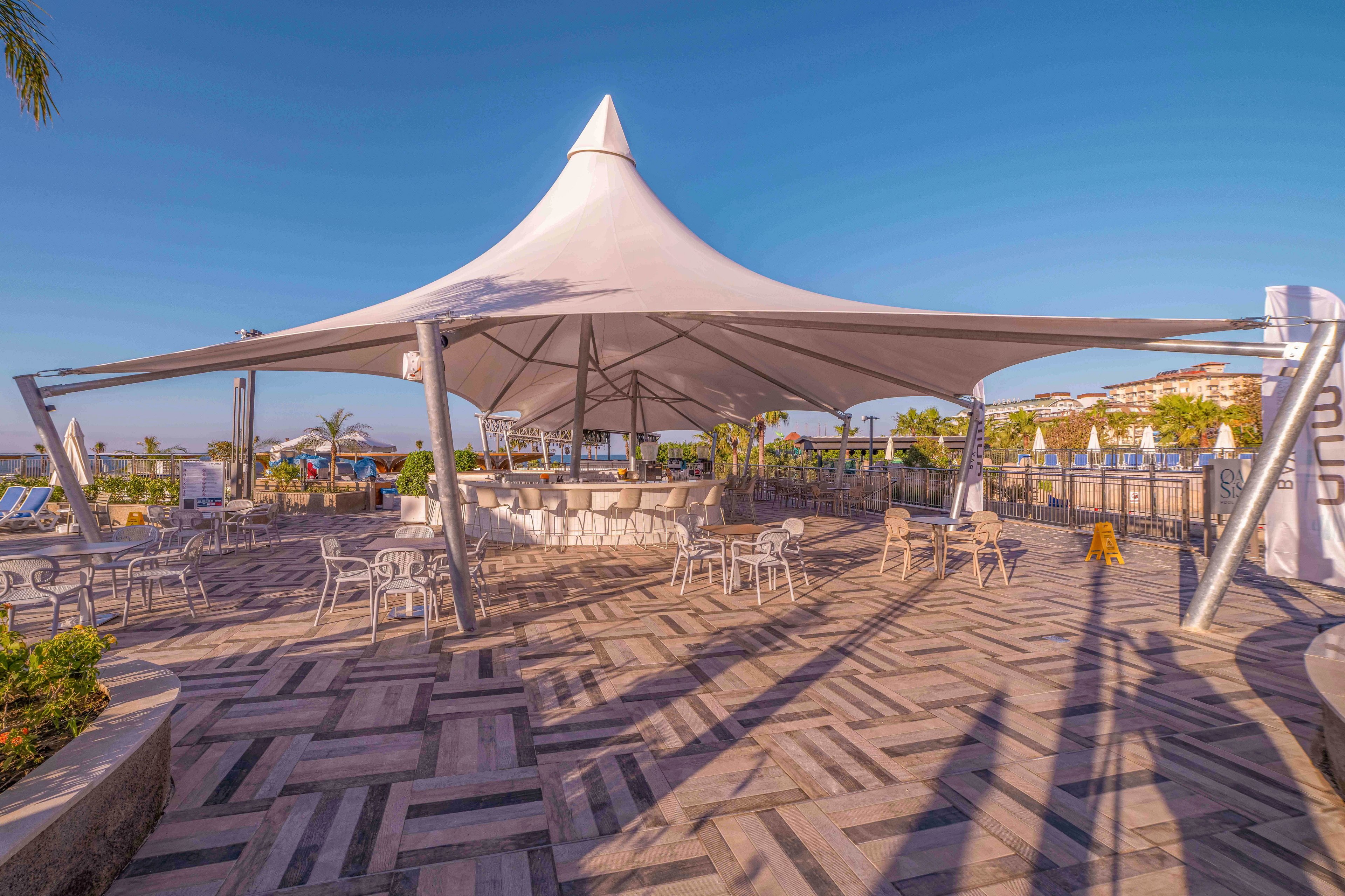 A spacious outdoor terrace features a large white tensile canopy structure at its center. Empty tables and chairs are arranged beneath the shade, suggesting a relaxed café or lounge setting. The patterned stone floor and surrounding palm trees create a resort-like atmosphere under a clear blue sky. Warm sunlight and long shadows add a tranquil, late-afternoon mood to the scene.