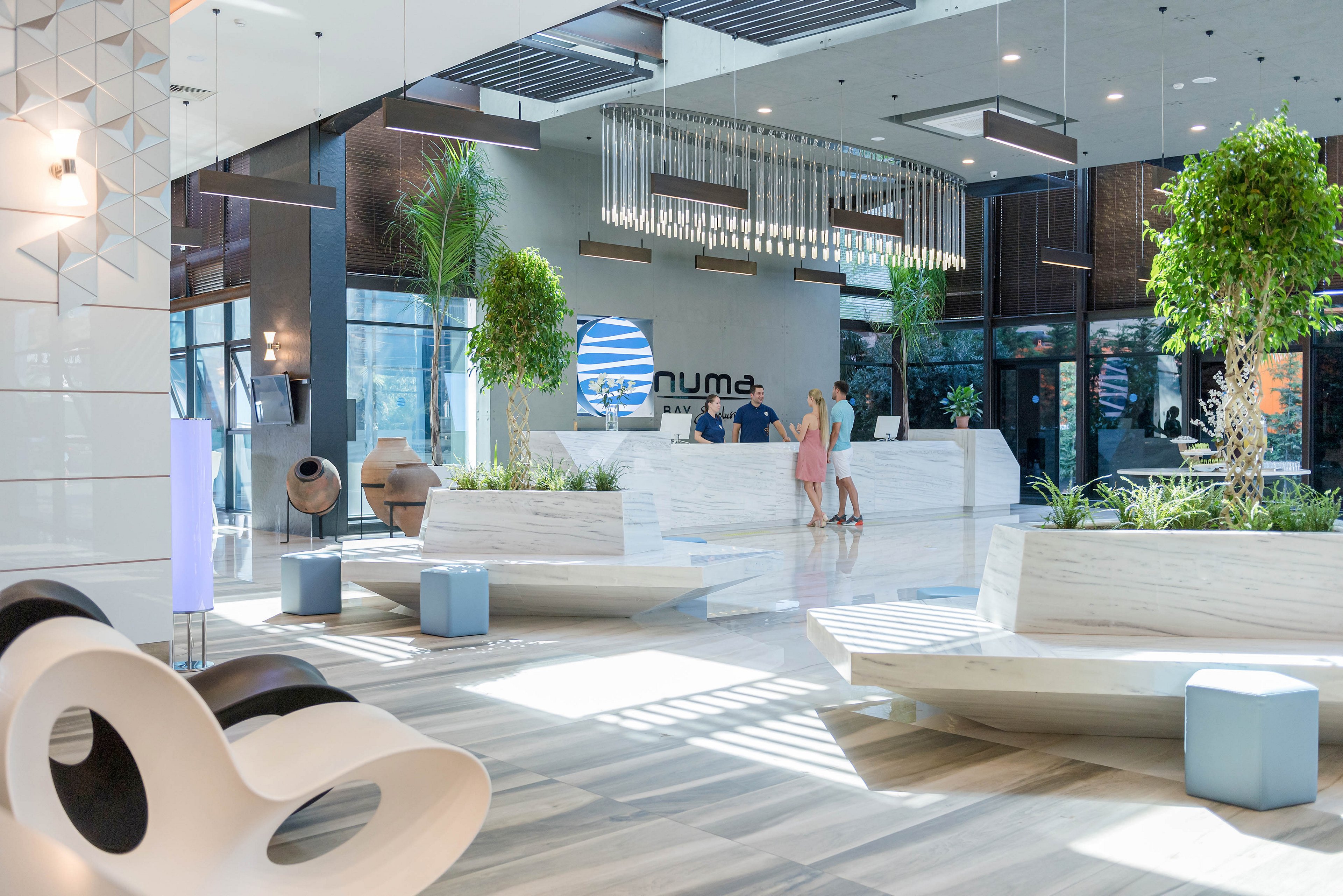 A spacious, contemporary lobby features a sleek white reception desk where two staff members assist a guest. Large windows and polished floors create a bright, airy atmosphere with abundant natural light. Modern seating, indoor plants, and geometric decor elements add to the stylish design. The scene conveys a welcoming, upscale environment suitable for hospitality or corporate settings.