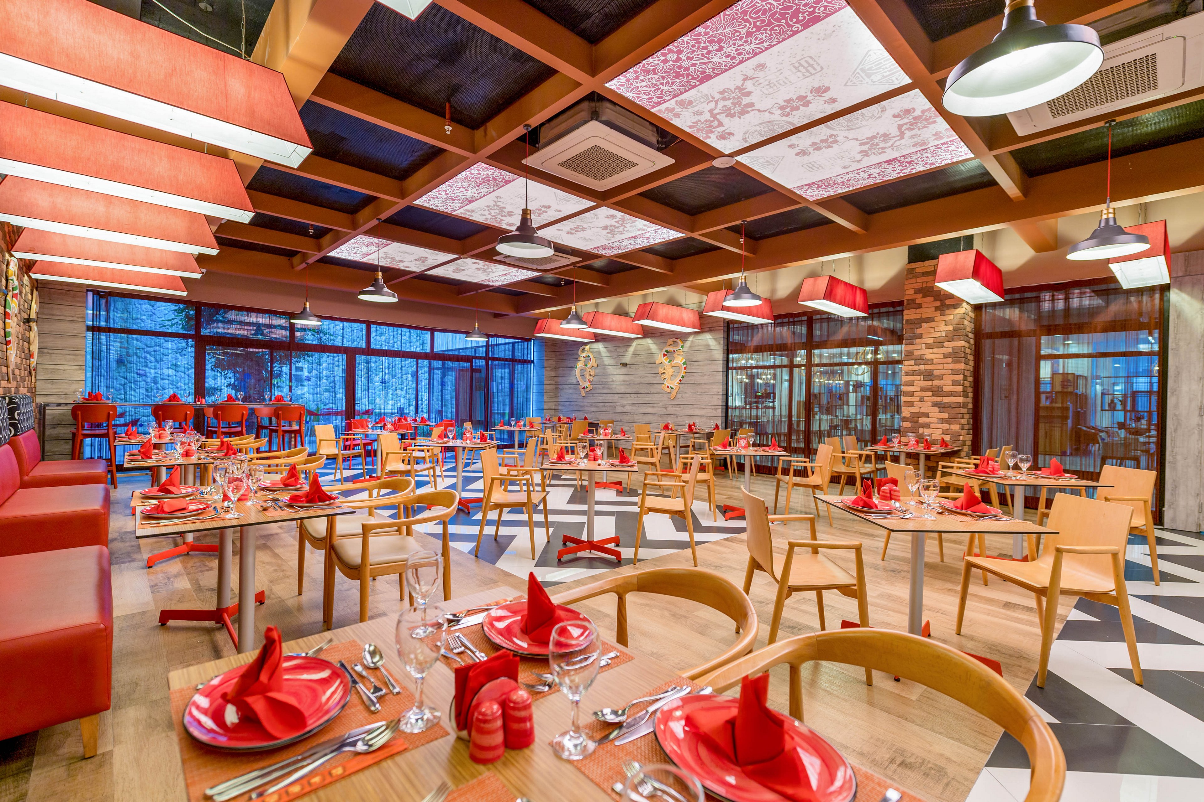 Spacious contemporary restaurant dining room with neatly arranged wooden tables and chairs. Each table is set with red plates, folded red napkins, cutlery, and glassware, creating a coordinated look. Large windows and warm ceiling lights illuminate the space, highlighting decorative wall elements and patterned ceiling panels. The overall atmosphere feels inviting, organized, and ready for guests.