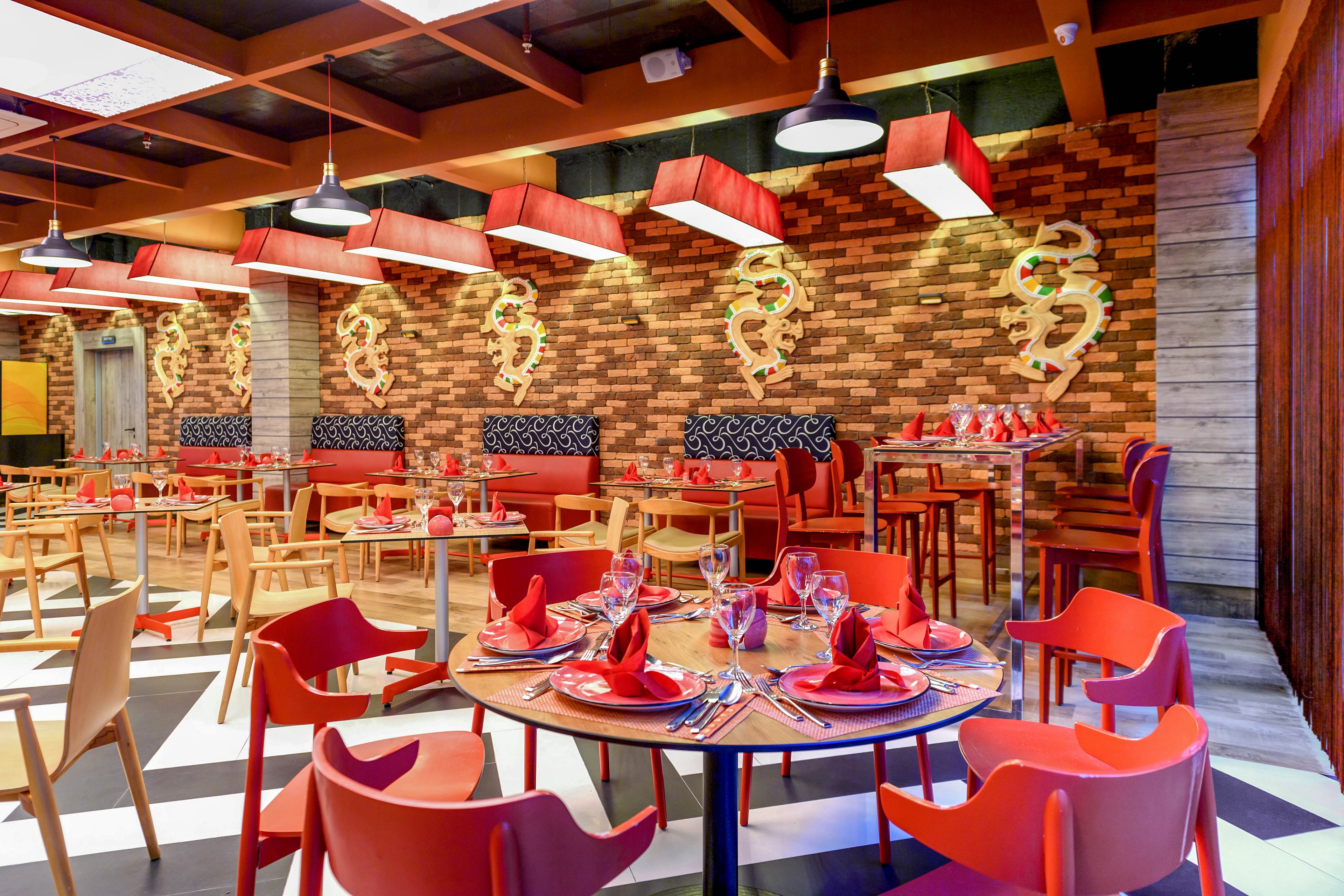 A vibrant restaurant dining area features round and square tables set with red napkins, glassware, and cutlery. The space has a modern design with red chairs, patterned bench seating, and a checkered black-and-white floor. Warm lighting hangs from a wooden ceiling grid, illuminating decorative wall art with swirling shapes on a brick backdrop. The overall atmosphere is contemporary, inviting, and ready for guests.