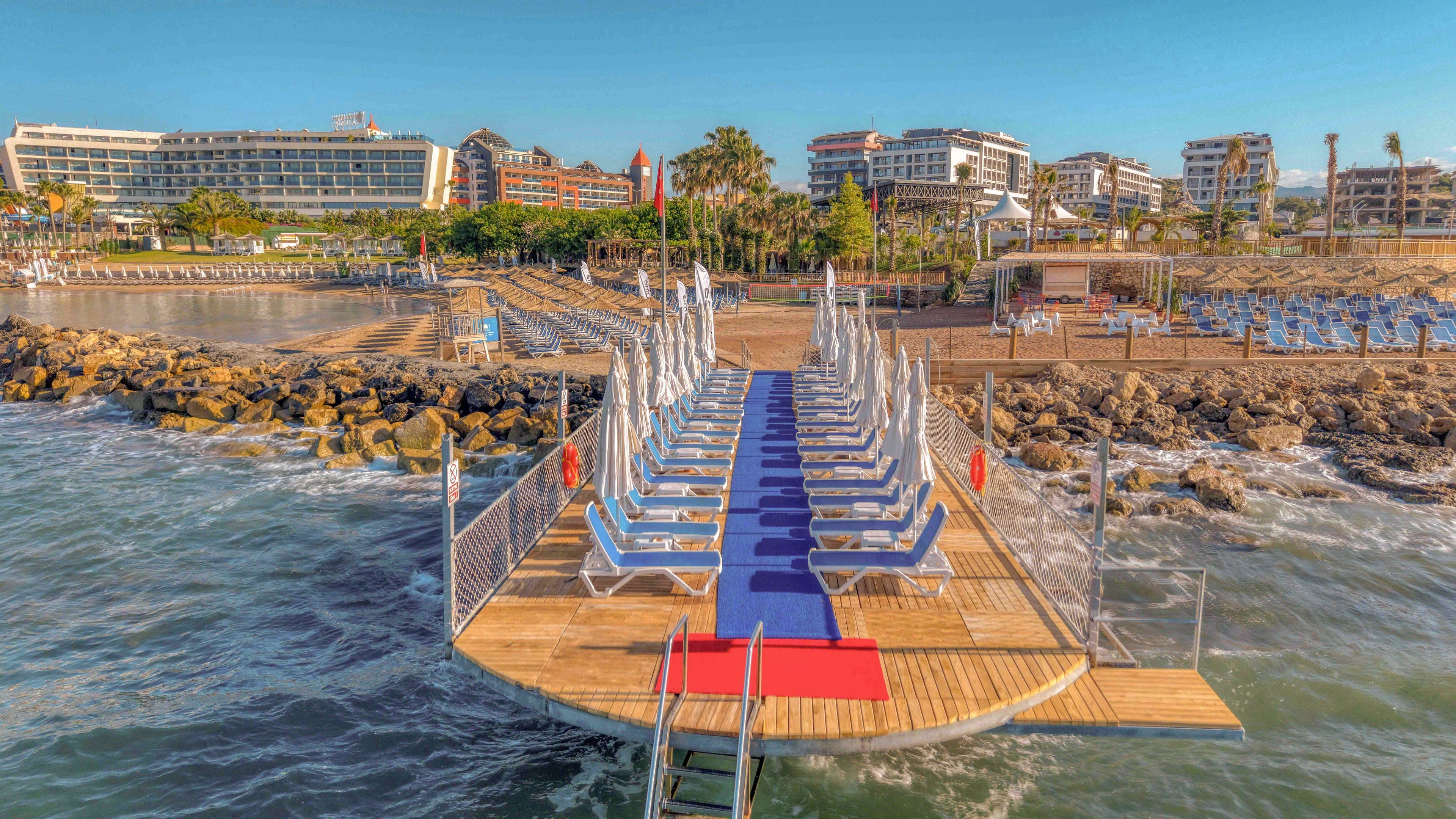 A wooden pier extends over the sea, lined with neatly arranged sun loungers and parasols facing the water. A blue carpet runs down the center of the pier, with a small red section near the ladder leading into the ocean. Rocky breakwaters and a sandy beachfront with additional seating areas are visible in the background. Modern resort buildings and palm trees frame the coastline under a clear, sunny sky.