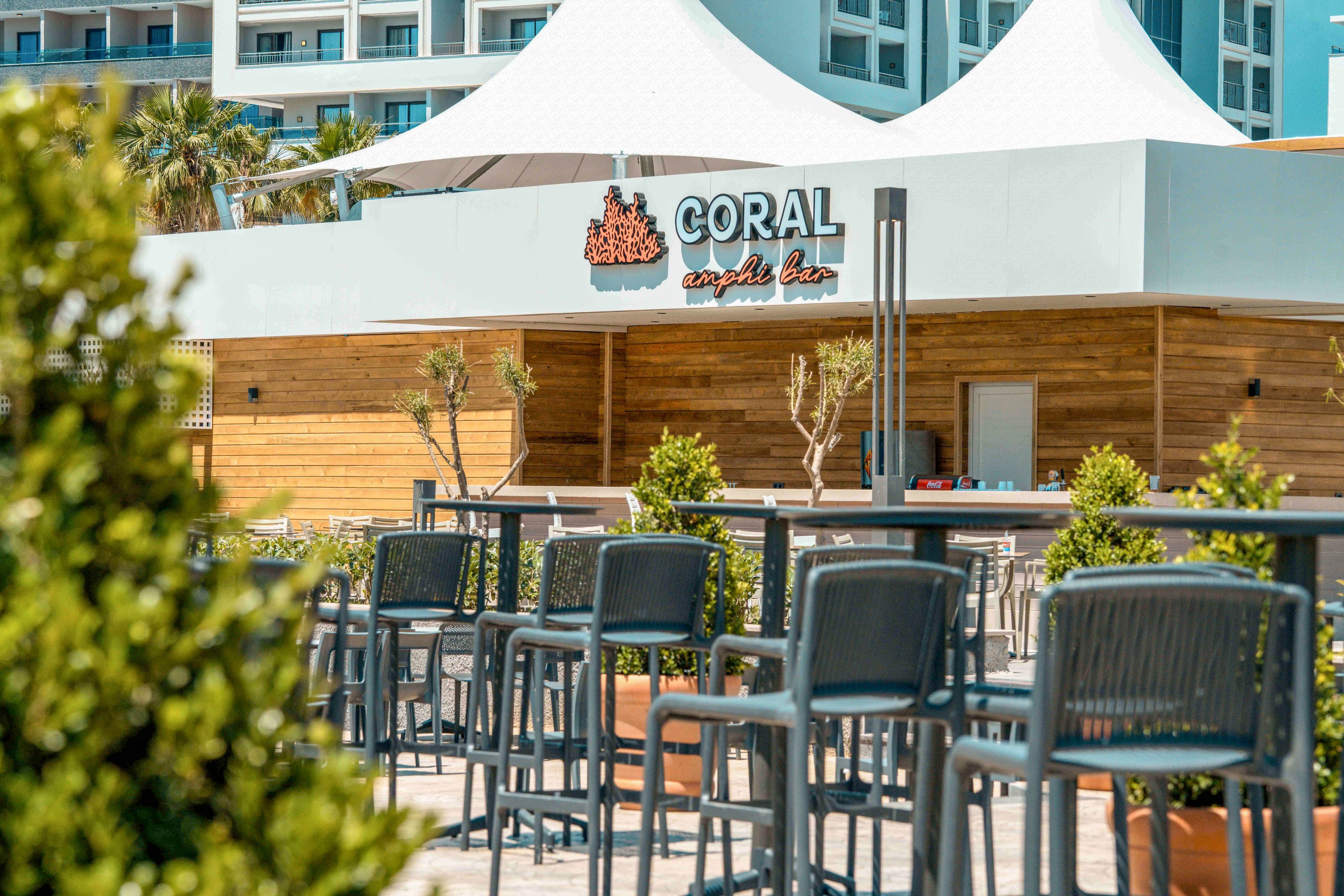 An outdoor bar named Coral Seaside Bar features rows of modern high chairs and tables on a sunlit terrace. The setting appears to be part of a resort complex, with contemporary buildings and white canopy structures in the background. Potted plants and small trees add greenery and a relaxed atmosphere to the wooden deck area. The scene suggests a casual, coastal venue for drinks and socializing in warm weather.