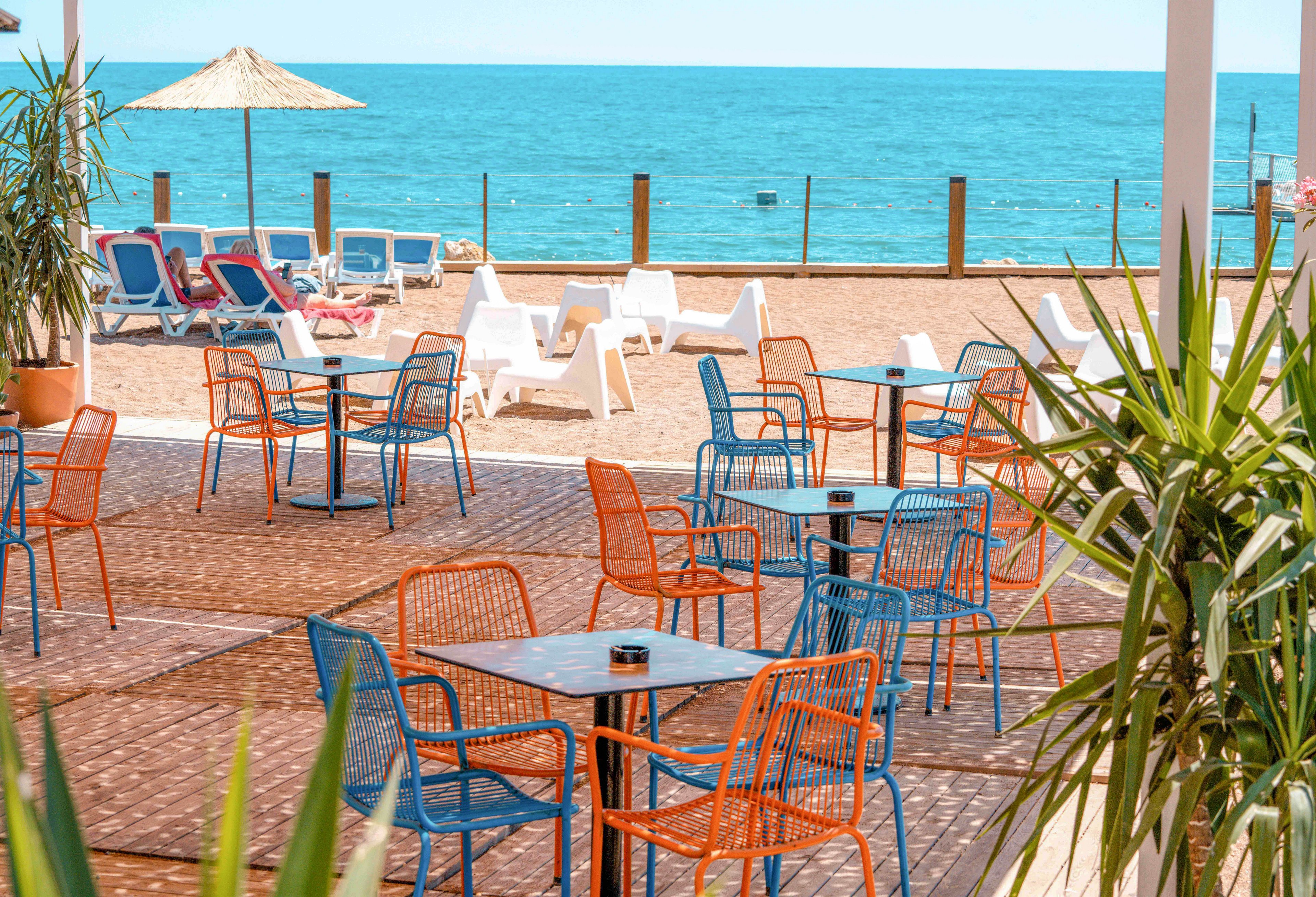 Outdoor café tables and chairs sit on a sunlit terrace overlooking a sandy beach and bright blue sea. Vibrant orange and blue metal chairs surround dark square tables, creating a lively and modern atmosphere. In the background, empty sun loungers and a straw parasol line the shore, suggesting a relaxed holiday setting. Green potted plants frame the scene, adding a touch of nature to the coastal environment.