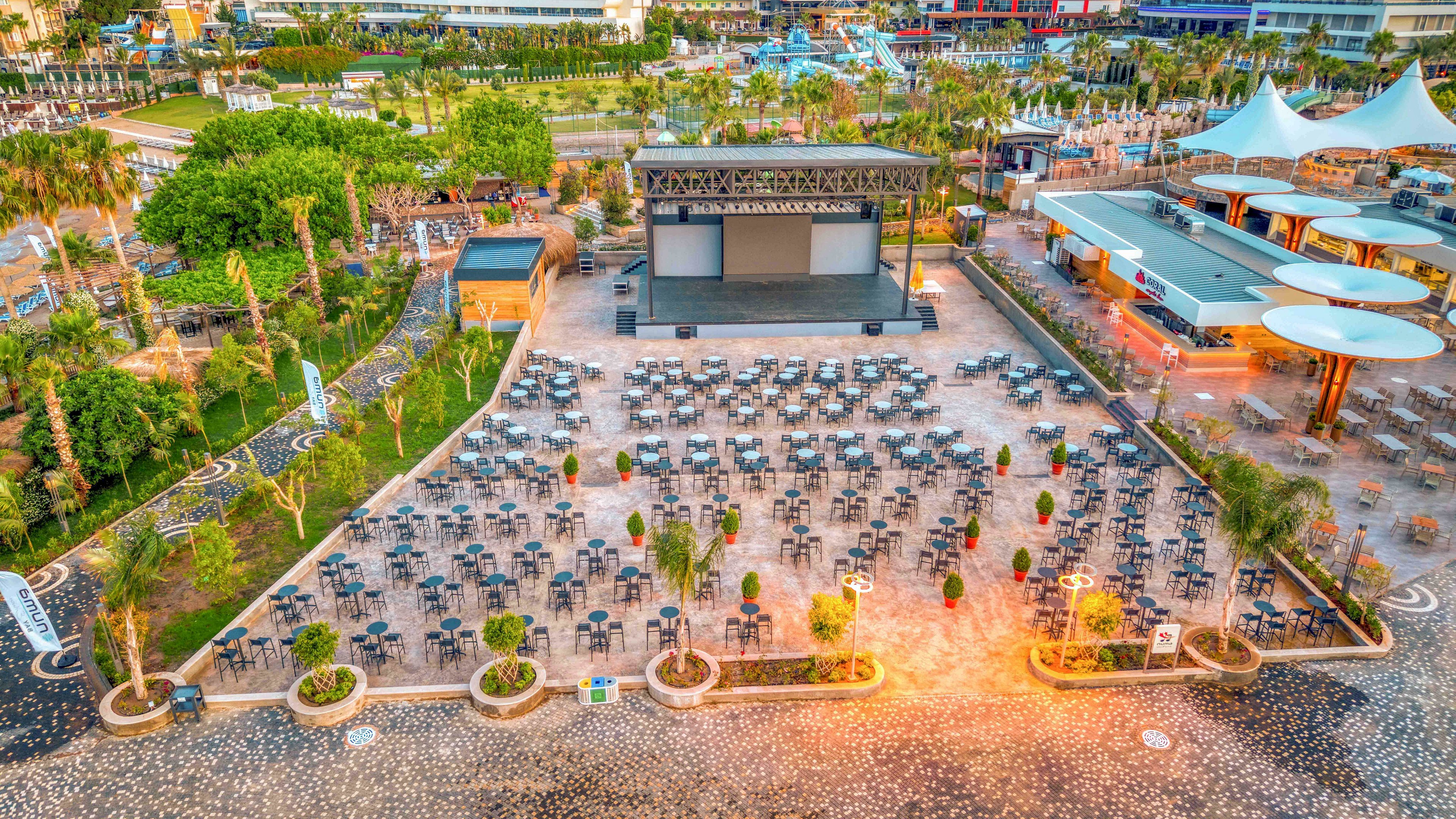 A large outdoor event venue features a central stage with a wide screen and rows of empty chairs arranged in front. The setting appears to be part of a resort or entertainment complex, surrounded by palm trees, landscaped greenery, and nearby buildings. Warm lighting illuminates the seating area and pathways, contrasting with the cooler tones of the surrounding structures. The scene suggests preparation for a concert, performance, or open-air presentation in a modern leisure environment.