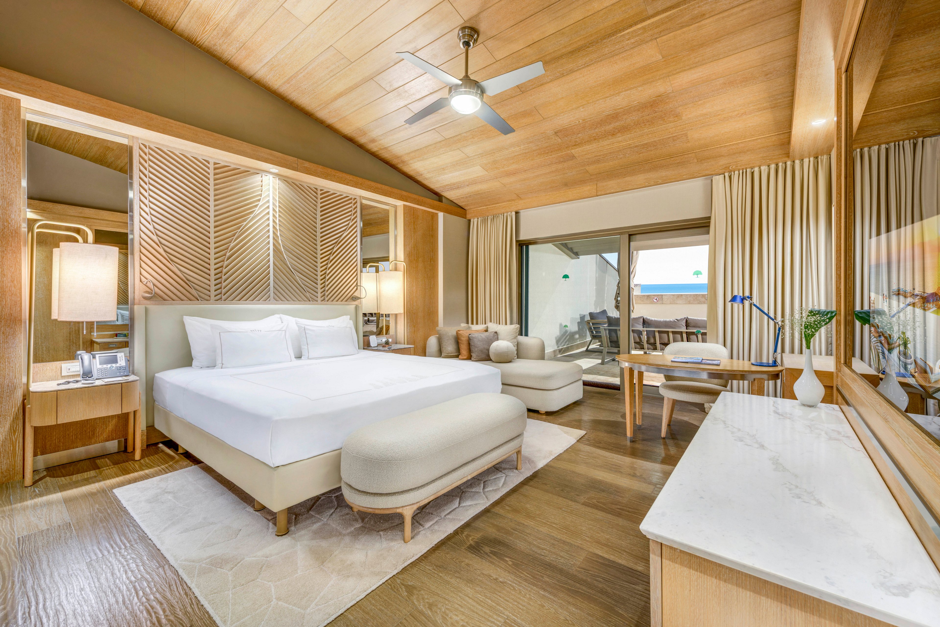 Spacious contemporary hotel room featuring a large bed with white linens and upholstered bench. The space includes wooden furniture, a ceiling fan, and warm ambient lighting. A sliding glass door opens to a terrace with outdoor seating and a partial ocean view. Neutral tones, light wood, and soft textiles create a calm, upscale atmosphere.