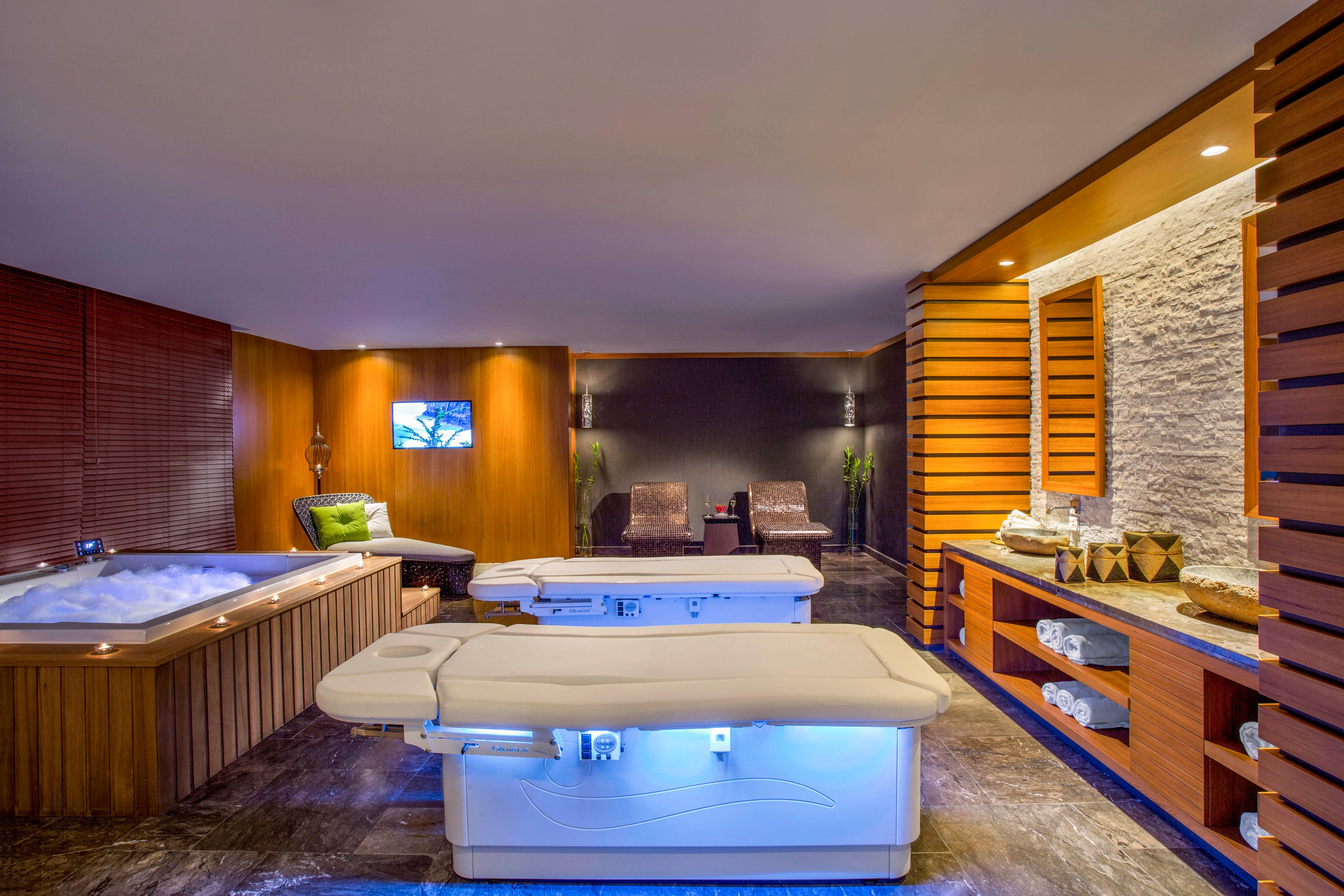 A modern spa treatment room features a central massage table illuminated with soft blue lighting. To the left, a bubbling jacuzzi tub is built into a wooden surround, adding to the sense of relaxation. In the background, two lounge chairs sit against a dark accent wall with a small television mounted above. Warm wood paneling, stacked stone, and neatly rolled white towels create a serene, upscale atmosphere.