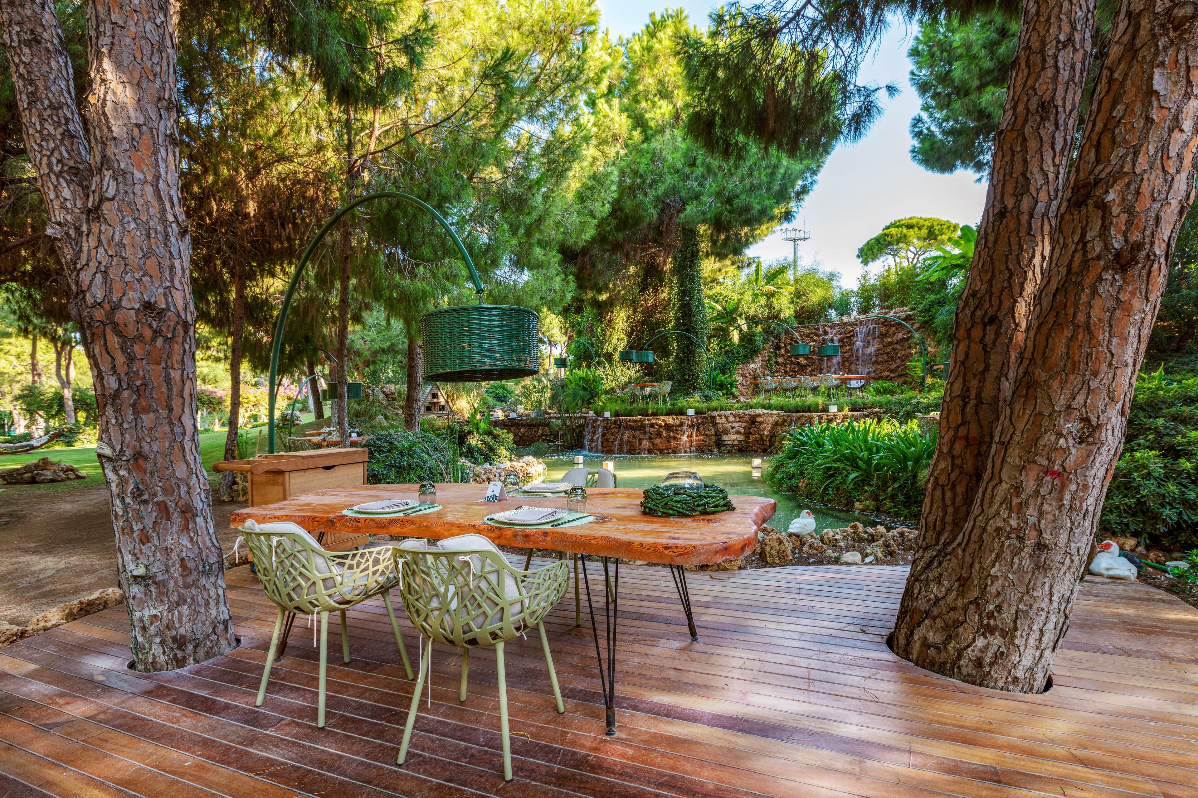 A wooden dining table is set for four on a polished deck between tall pine trees. Modern chairs surround the table, which is arranged with plates and decorative centerpieces. In the background, a lush garden with manicured greenery and a small pond creates a tranquil atmosphere. Soft natural light filters through the trees, enhancing the serene outdoor setting.