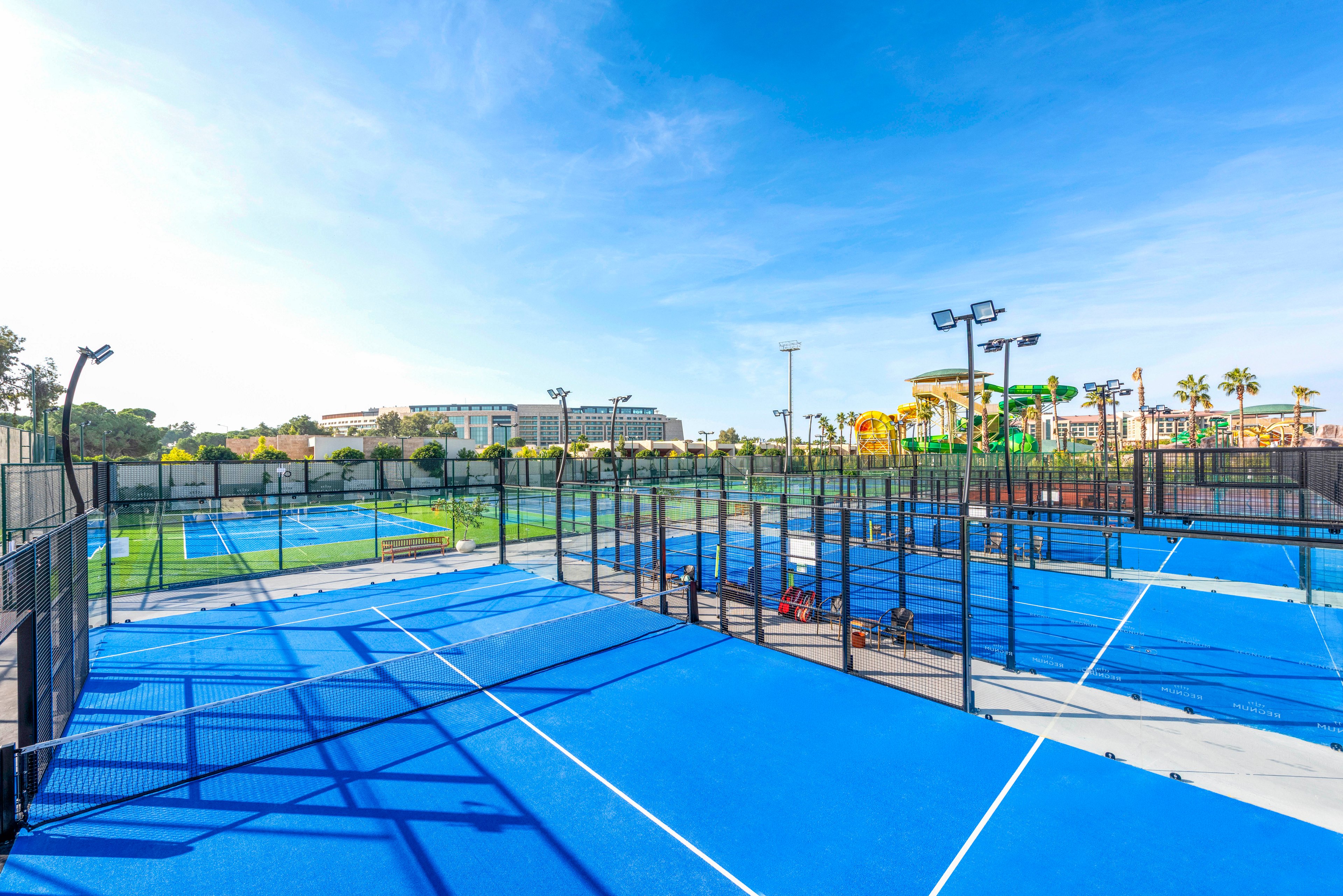 Multiple bright blue padel courts are arranged side by side in an outdoor sports facility. The courts are surrounded by tall black metal fences and lighting poles under a clear blue sky. In the background, resort-style buildings, palm trees, and a colorful water park area are visible. The scene suggests a sunny vacation destination with modern recreational amenities.