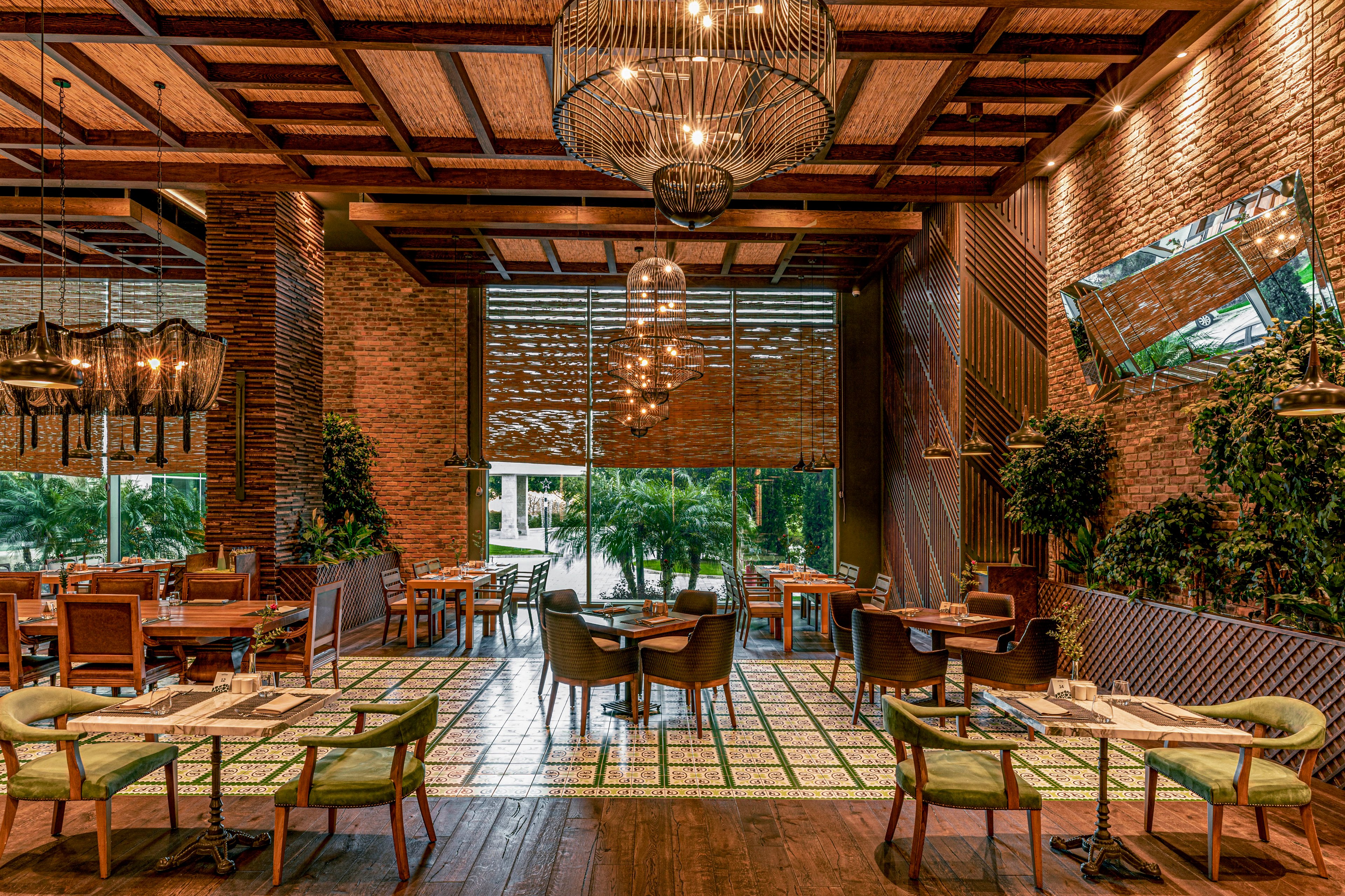 Upscale restaurant dining room featuring wooden tables and upholstered chairs is shown empty and ready for guests. The space combines exposed brick walls, patterned tile flooring, and large windows overlooking lush outdoor greenery. Decorative pendant lights and modern chandeliers hang from a wooden ceiling, creating a warm and inviting ambiance. Potted plants and vertical green accents enhance the natural, sophisticated atmosphere.