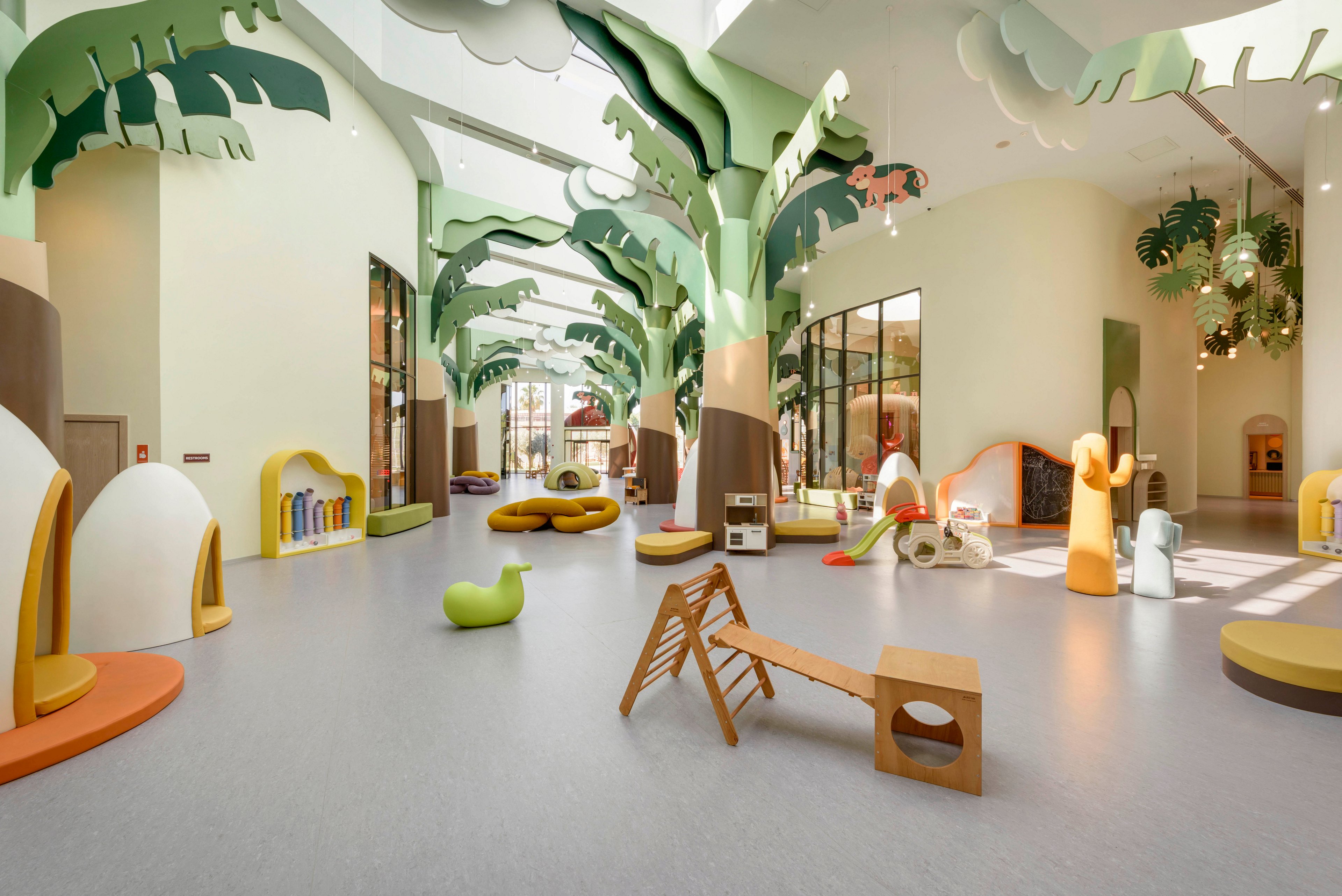 Spacious indoor children’s play area featuring whimsical, oversized tree structures and soft play equipment. The room is filled with sculptural seating, slides, and climbing elements in organic shapes. Large windows and high ceilings create an airy, light-filled environment. Soft pastel colors and rounded forms give the space a playful, imaginative atmosphere.