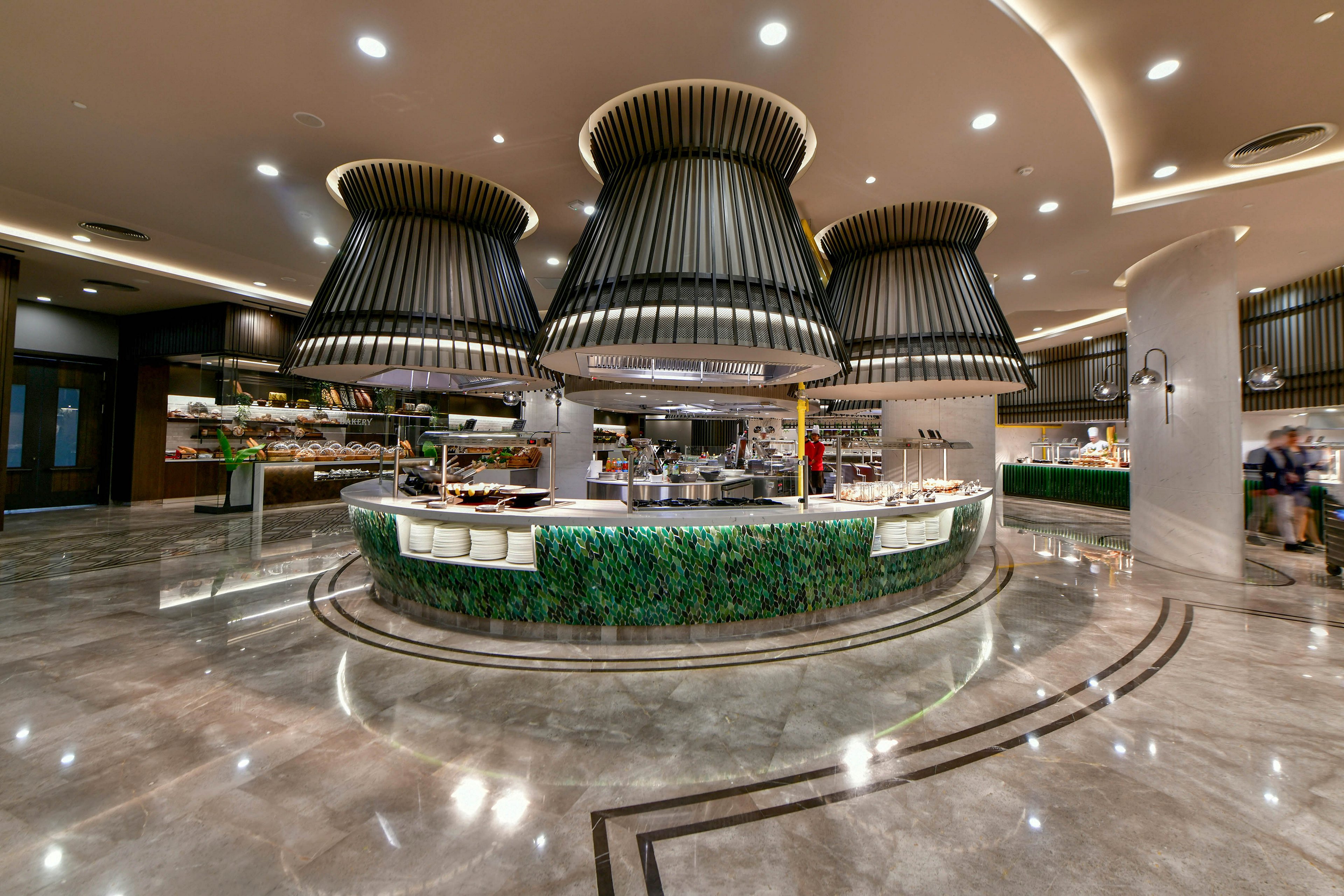 A spacious, modern buffet station stands in the center of a polished dining hall. Large sculptural pendant lights hang above the circular counter, drawing attention to the food display. The counter is wrapped in green foliage-like panels, contrasting with the glossy marble floor and neutral ceiling. The setting appears clean, upscale, and designed for a hotel or resort dining experience.
