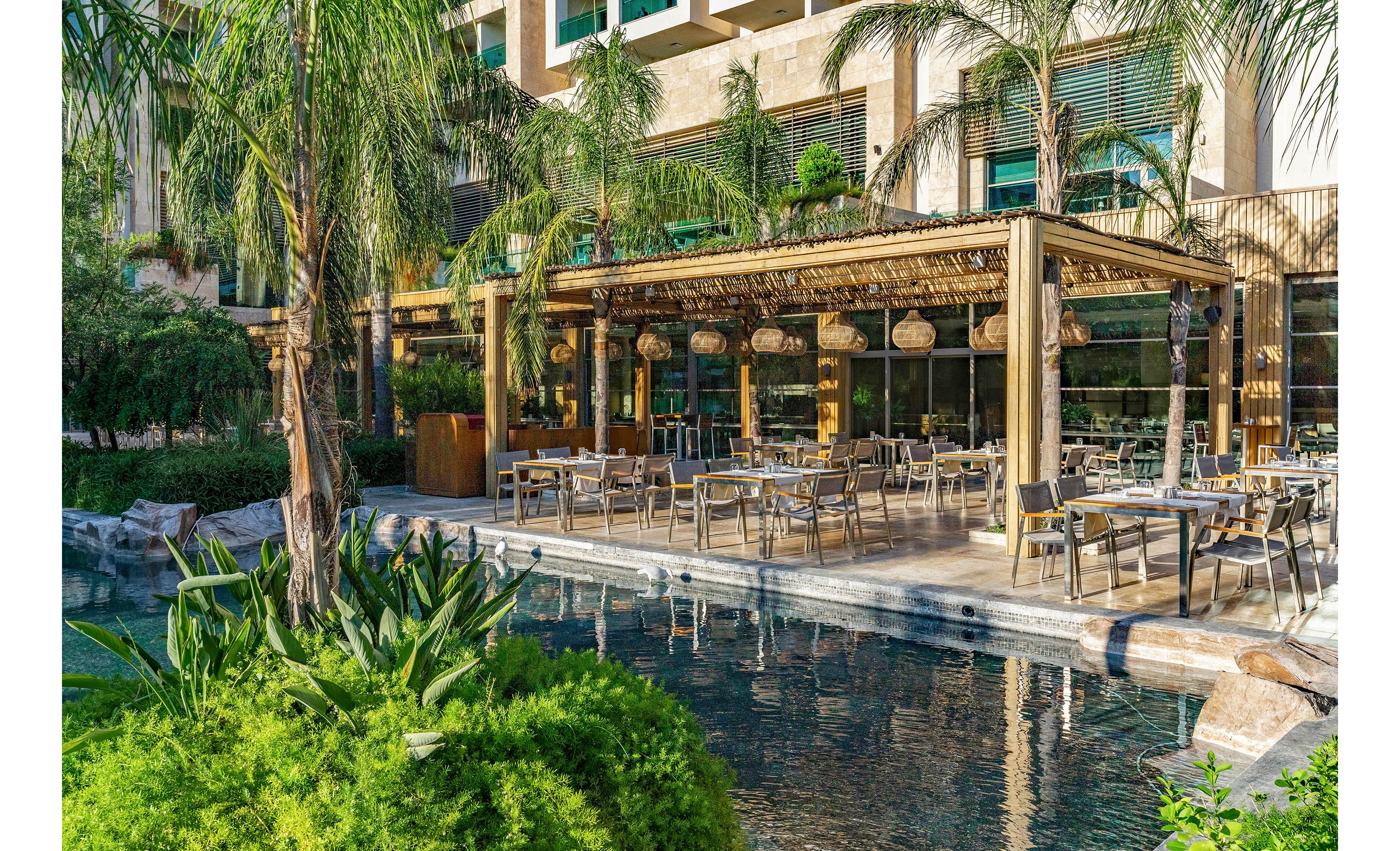 An open-air restaurant terrace is set beside a clear reflective water feature at a modern resort. Wooden pergolas with woven shades and hanging basket lights cover neatly arranged dining tables and chairs. Lush tropical plants and palm trees surround the seating area, creating a tranquil oasis atmosphere. Large windows of the adjacent building reflect the greenery and outdoor setting, emphasizing a relaxed vacation or leisure dining experience.
