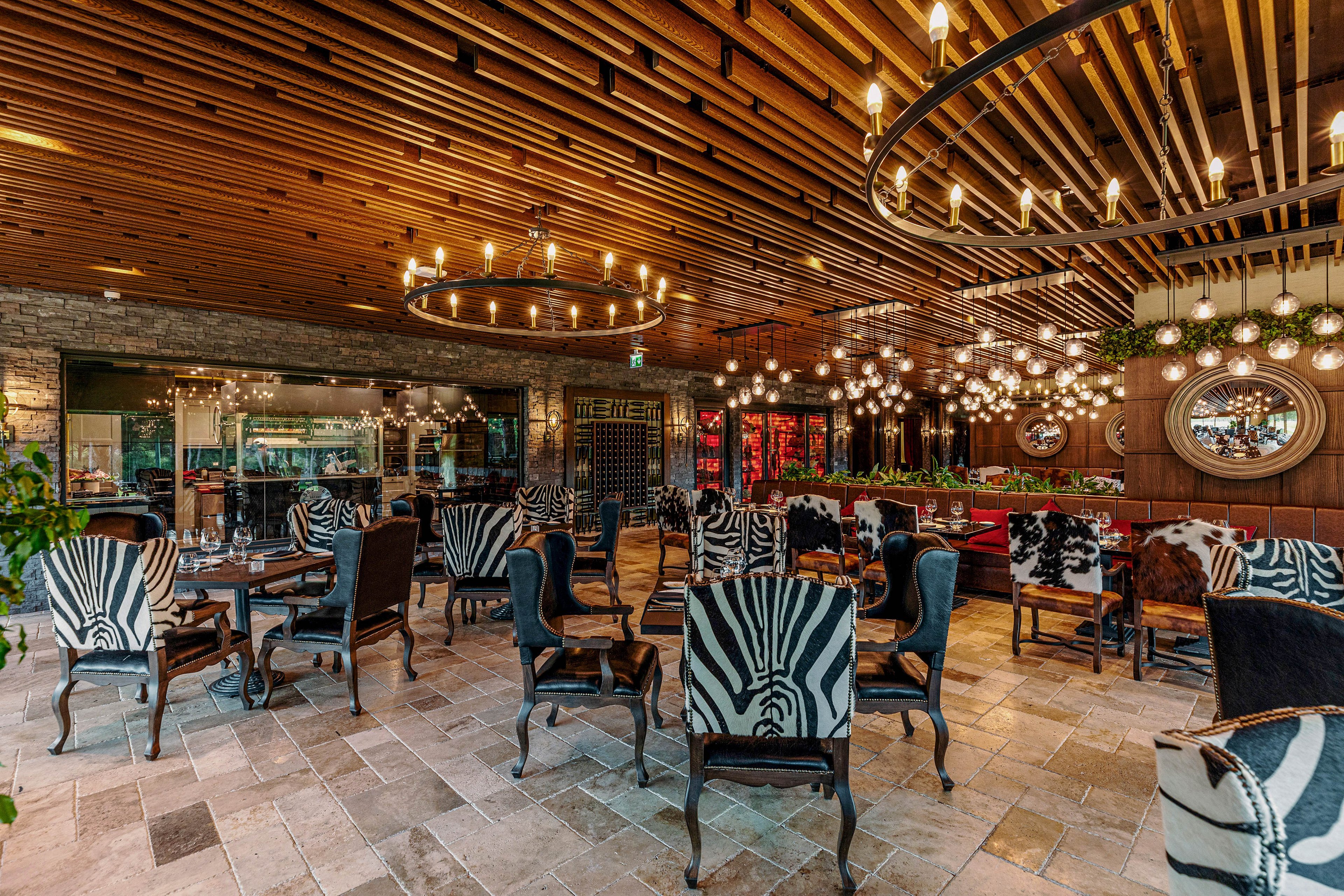 Upscale restaurant interior featuring distinctive zebra print chairs arranged around dark wooden tables. The space has a warm ambiance with numerous hanging light fixtures and circular chandeliers on a slatted wooden ceiling. Stone walls, large mirrors, and greenery accents add texture and depth to the dining area. The restaurant appears empty, highlighting the decor, layout, and inviting atmosphere.