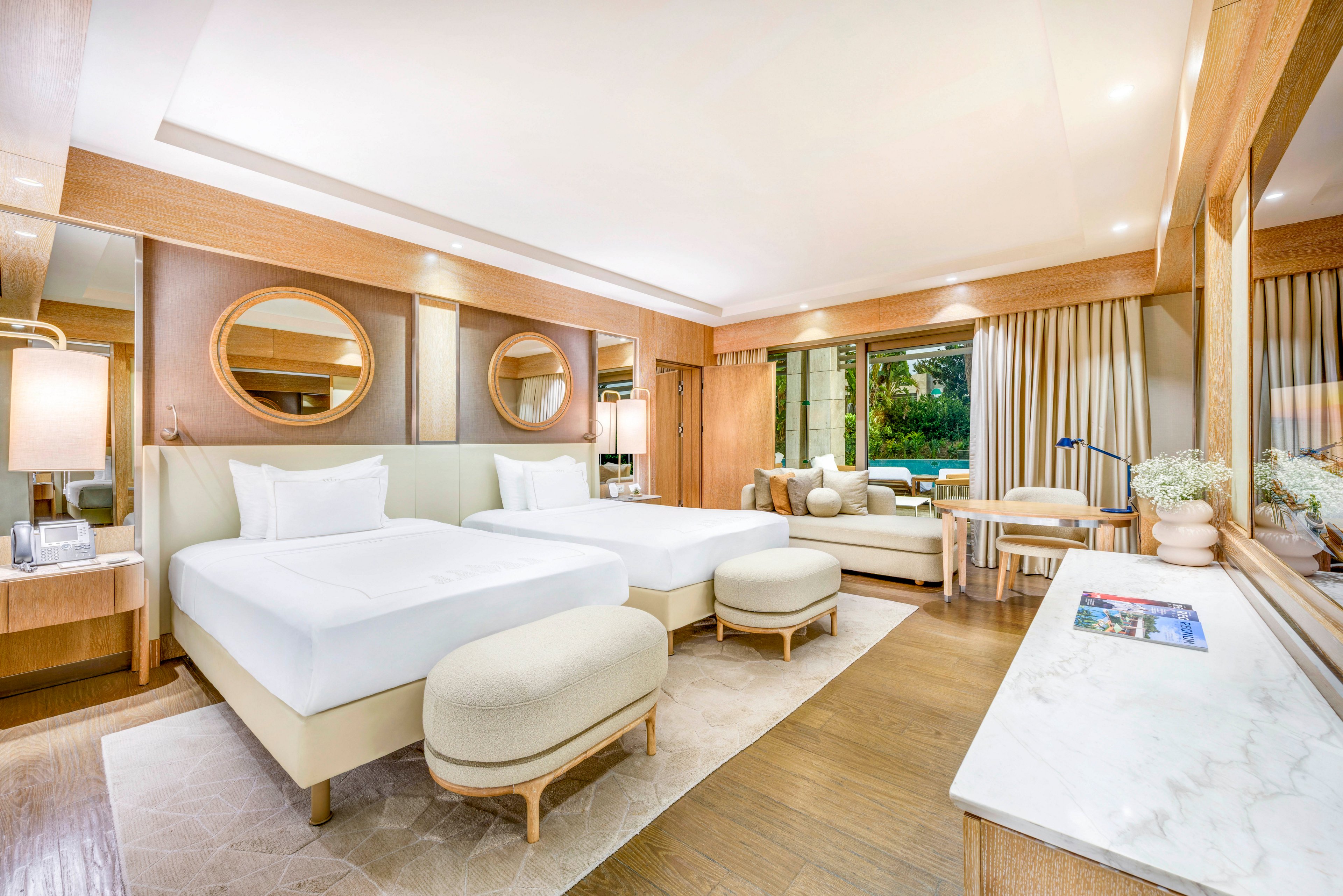 Spacious contemporary hotel room featuring two neatly made twin beds with white linens and upholstered headboards. The interior showcases light wood furniture, including bedside tables, a cushioned bench, and a marble-topped console. Large mirrors, soft lighting, and floor-to-ceiling curtains create an inviting, luxurious atmosphere. A seating area with a sofa and table overlooks greenery outside through wide windows.