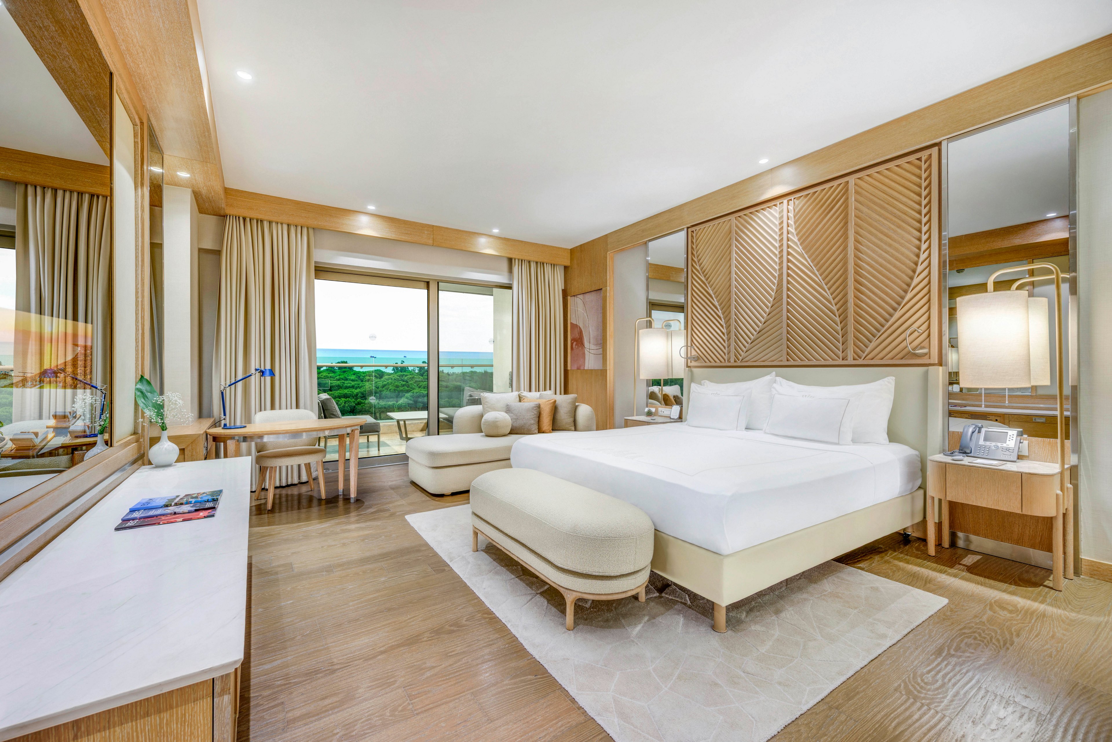Spacious contemporary hotel room featuring a large bed with crisp white linens and upholstered bench seating. The interior showcases light wood finishes, soft neutral tones, and floor-to-ceiling curtains. A desk and seating area sit near the sliding glass doors that open to a balcony overlooking greenery and the ocean. Recessed lighting and large mirrors enhance the bright, airy atmosphere of the space.