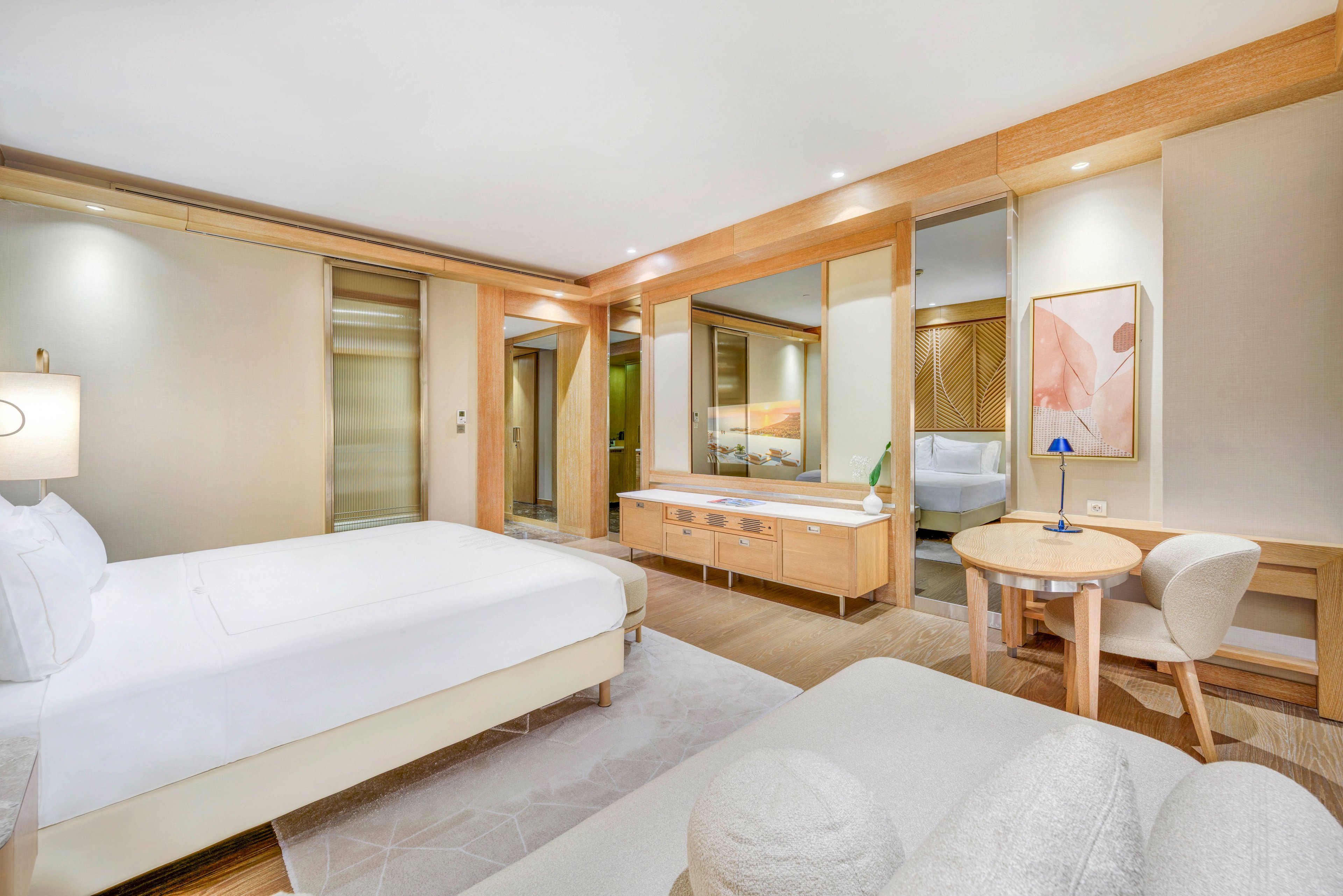 Spacious contemporary hotel room featuring a large neatly made bed and light wood furnishings. A built-in vanity with mirror and drawers lines one wall, reflecting the room and adding depth. A small round table with a chair and desk lamp creates a cozy workspace near framed abstract artwork. Soft neutral tones, recessed lighting, and minimal decor give the space a calm, luxurious atmosphere.