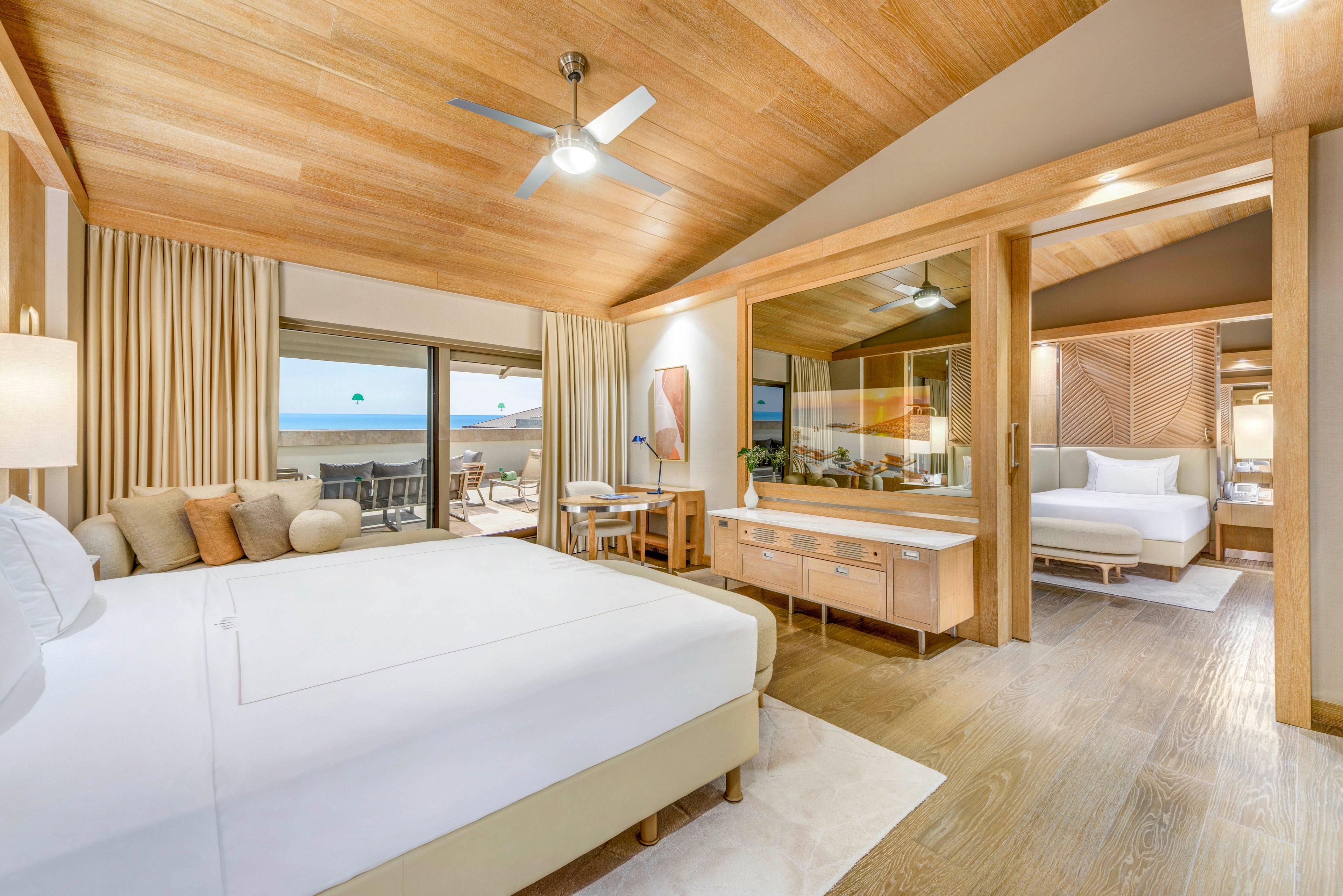Spacious contemporary hotel suite featuring two large beds and light wood finishes. The main room opens to a balcony with a clear view of the ocean and outdoor seating. Neutral tones, soft lighting, and natural textures create a calm, airy atmosphere. A glass partition separates the sleeping area from a modern bathroom vanity, enhancing the open-plan design.