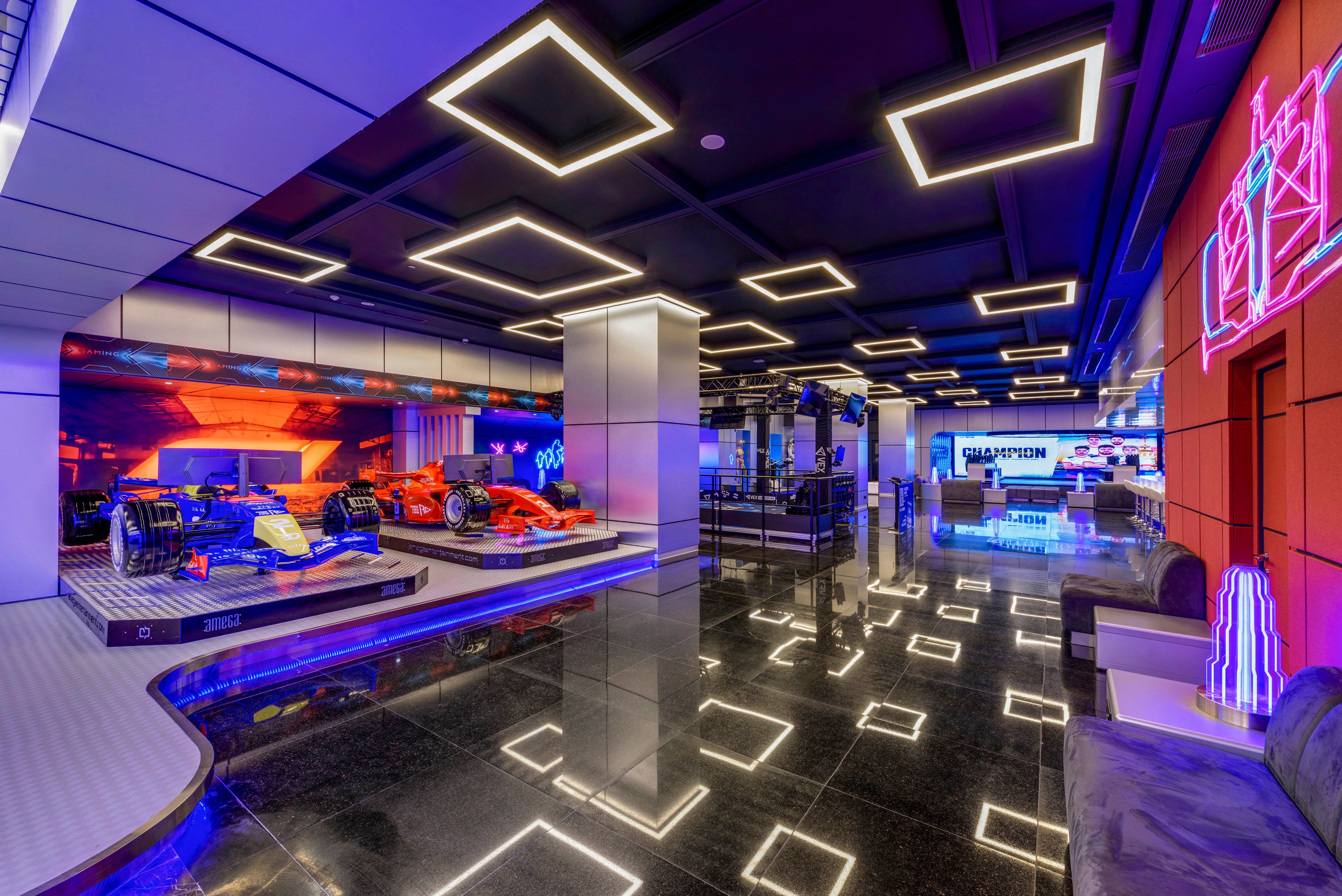 A modern indoor showroom features two Formula-style race cars displayed on raised platforms. The space has glossy black reflective flooring and illuminated geometric ceiling lights that create a high-tech atmosphere. Neon wall art and digital screens add vibrant color and visual interest throughout the room. Seating areas and counters in the background suggest an experiential exhibition or entertainment venue.