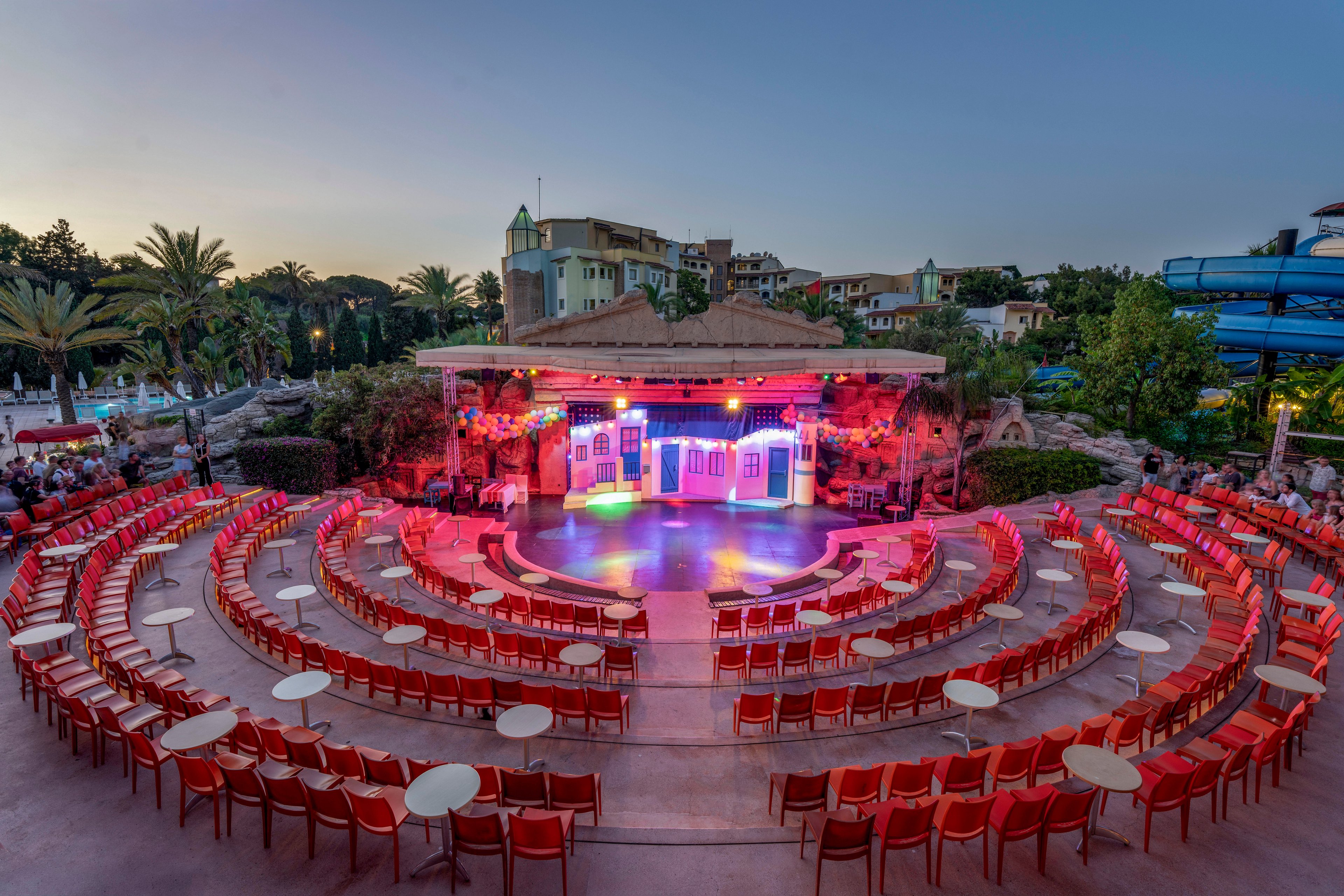 An outdoor amphitheater features concentric rows of red chairs and small round tables facing a brightly lit stage. The stage is decorated with colorful house-like facades and vibrant neon lighting, creating a festive atmosphere. Surrounding the venue are palm trees, lush greenery, and resort-style buildings under a clear dusk sky. A blue waterslide structure is visible to the right, suggesting a vacation or resort entertainment setting.