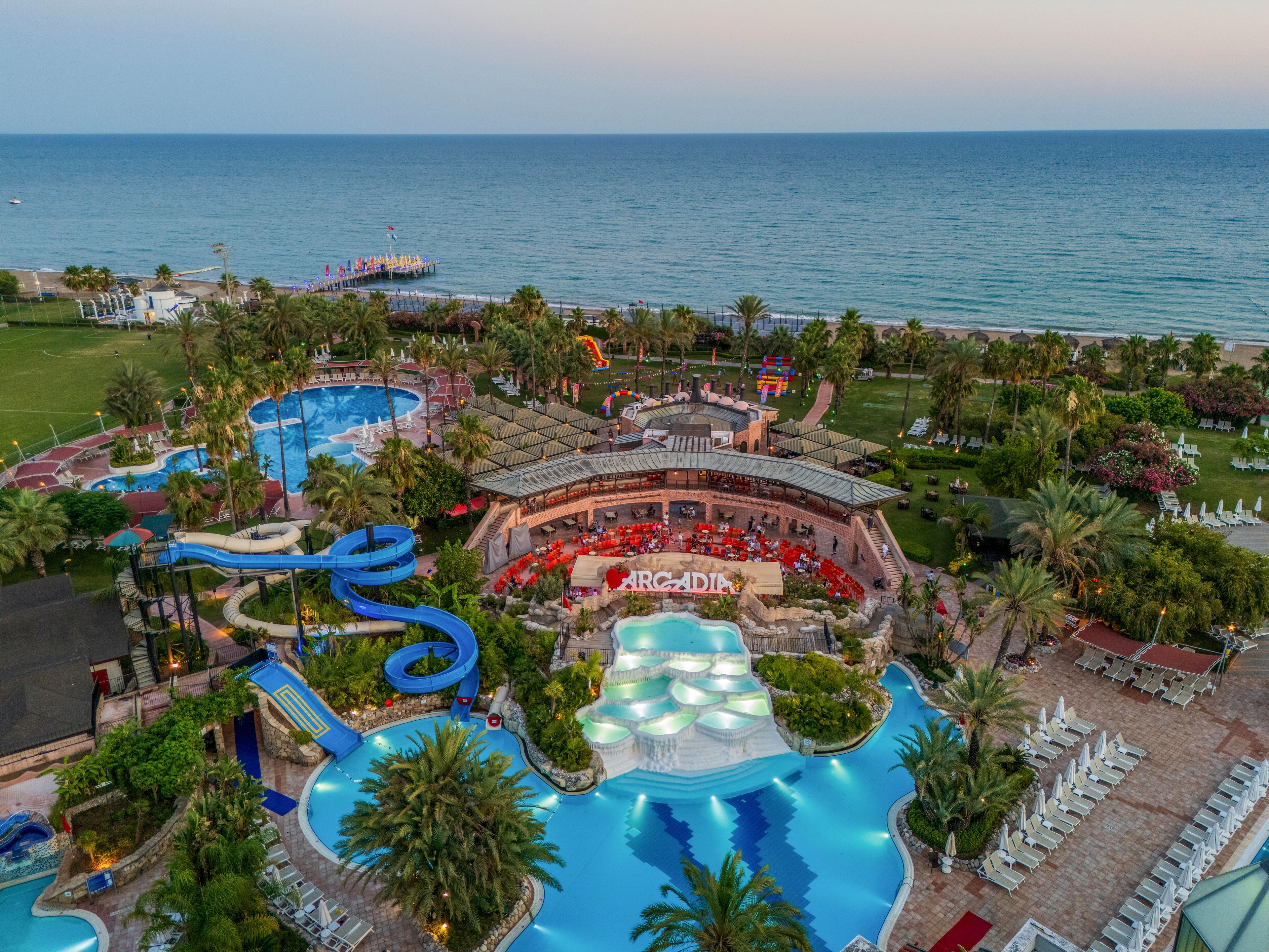 A large coastal resort complex features multiple outdoor swimming pools and winding blue water slides near the shoreline. The central area includes a circular bar or restaurant with the word "ARCADIA" illuminated above it, surrounded by palm trees and lush greenery. Sun loungers line the pool edges, and guests can be seen enjoying the facilities in the warm evening light. In the background, the calm sea stretches to the horizon, enhancing the vacation atmosphere.