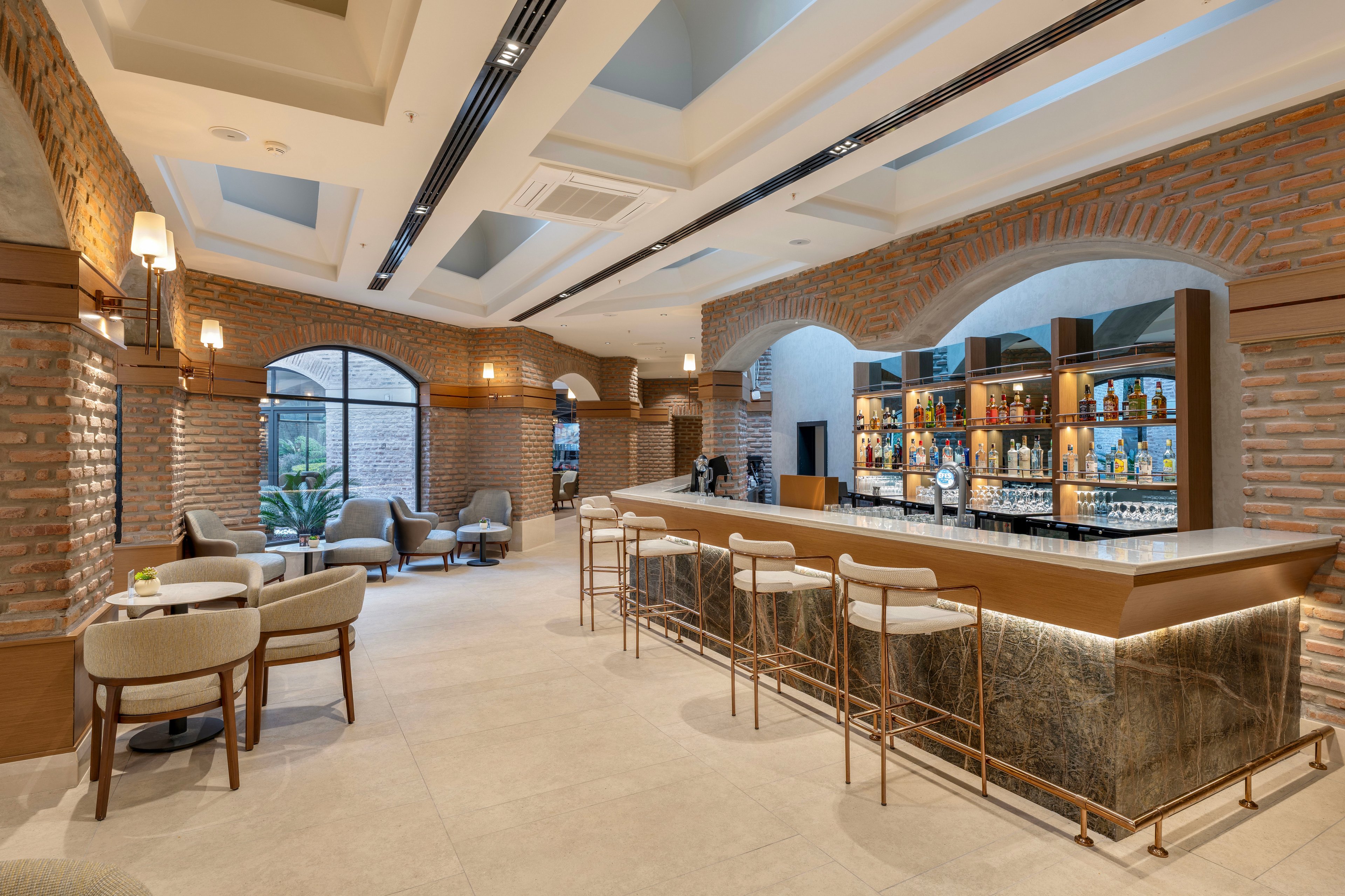 Contemporary bar interior featuring a long counter with high stools and a backlit liquor display. The space is framed by exposed brick arches, large windows, and recessed ceiling lighting. Comfortable lounge chairs and small tables create a relaxed seating area near the windows. The overall atmosphere is warm and inviting, suitable for socializing or casual meetings.