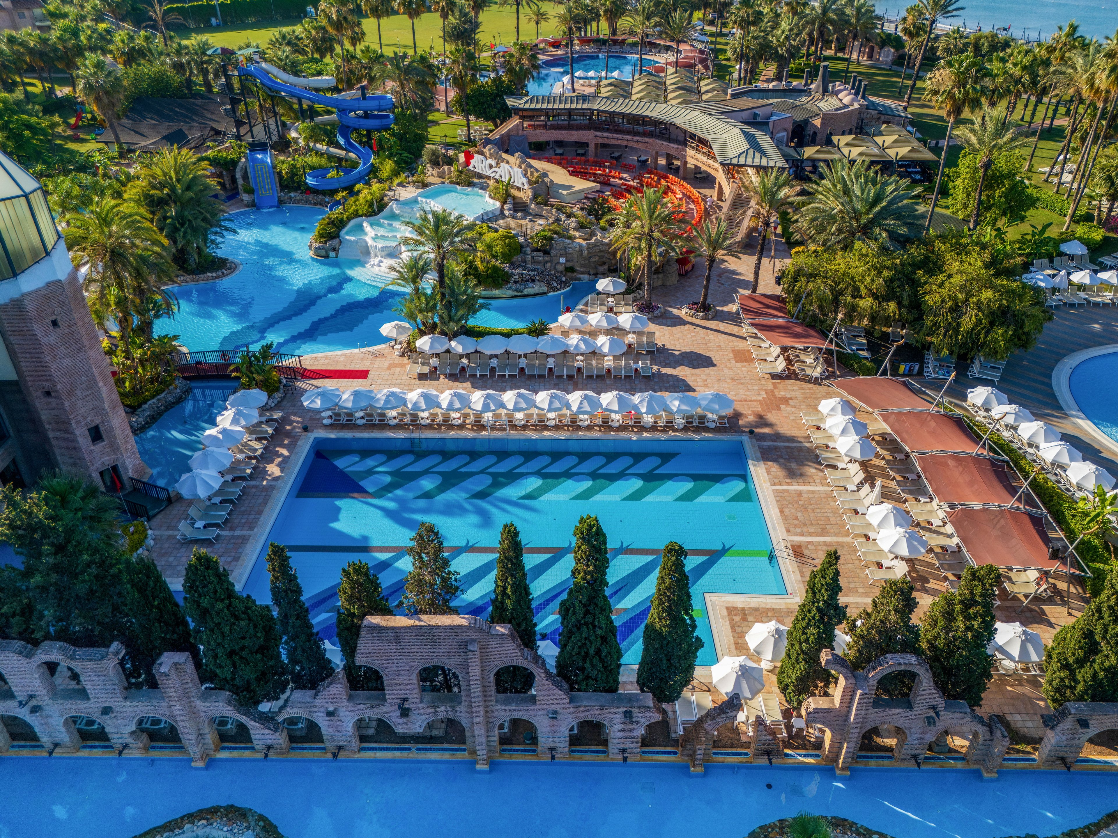 A large outdoor swimming pool complex is captured from above at a luxury resort. Multiple pools of varying shapes are surrounded by rows of white sun loungers and umbrellas. Tall palm trees and manicured greenery frame the water areas, with slides and curved walkways visible in the background. The scene suggests a sunny vacation destination focused on relaxation and family leisure.