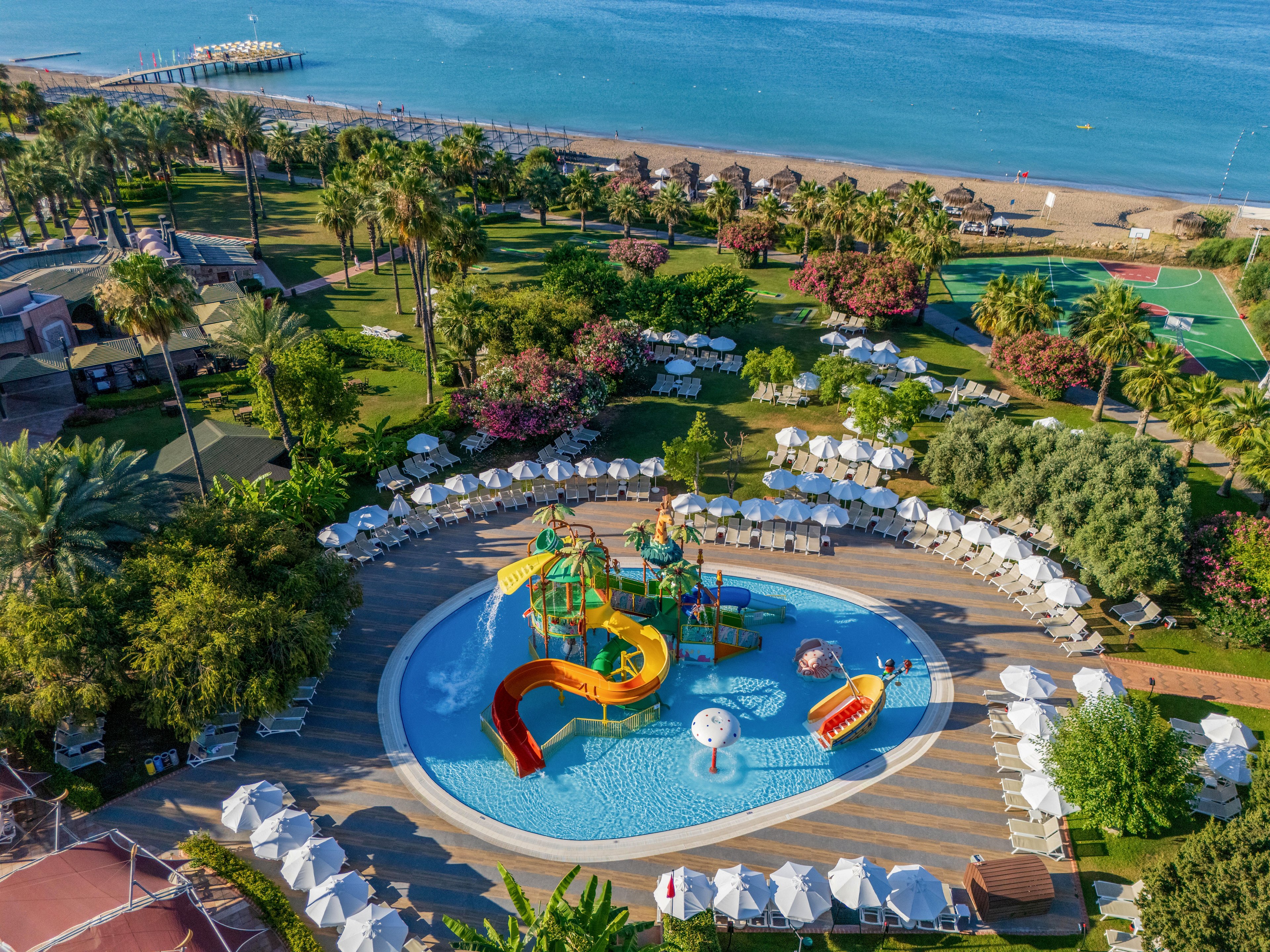 A colorful children’s water playground with multiple slides sits at the center of a resort pool. White sun loungers and umbrellas surround the pool in neat rows, creating a relaxed vacation atmosphere. Lush palm trees, gardens, and nearby sports courts extend toward a sandy beach and calm blue sea in the background. The scene suggests a family-friendly holiday destination with ample outdoor recreation.
