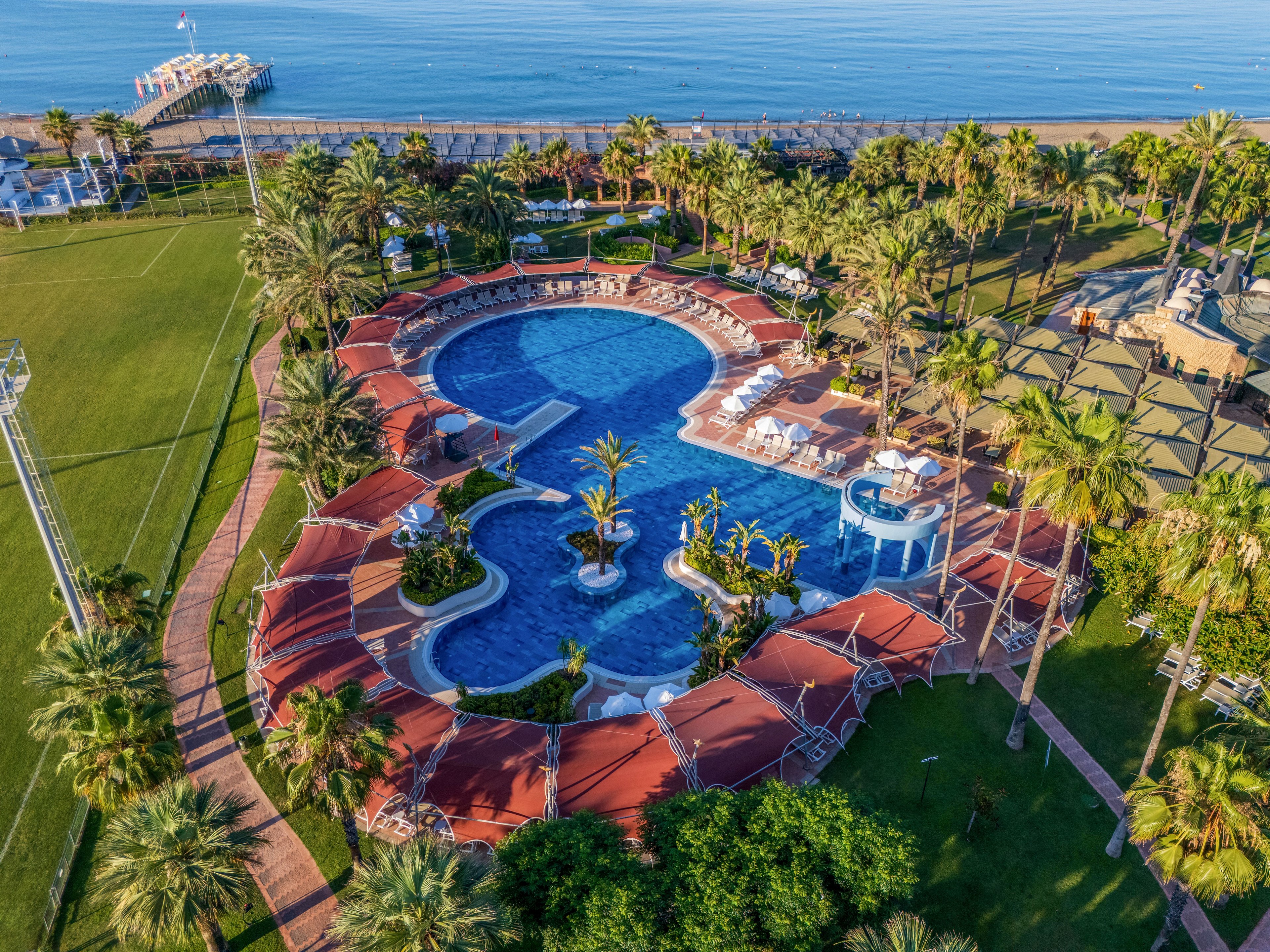 A large resort swimming pool is seen from above, surrounded by palm trees and red-roofed cabanas. Curved walkways and sun loungers frame the pool area, creating an inviting leisure setting. Beyond the pool, a green lawn leads toward the calm sea and a pier extending over the water. The overall scene suggests a sunny, tropical vacation destination focused on relaxation and outdoor recreation.