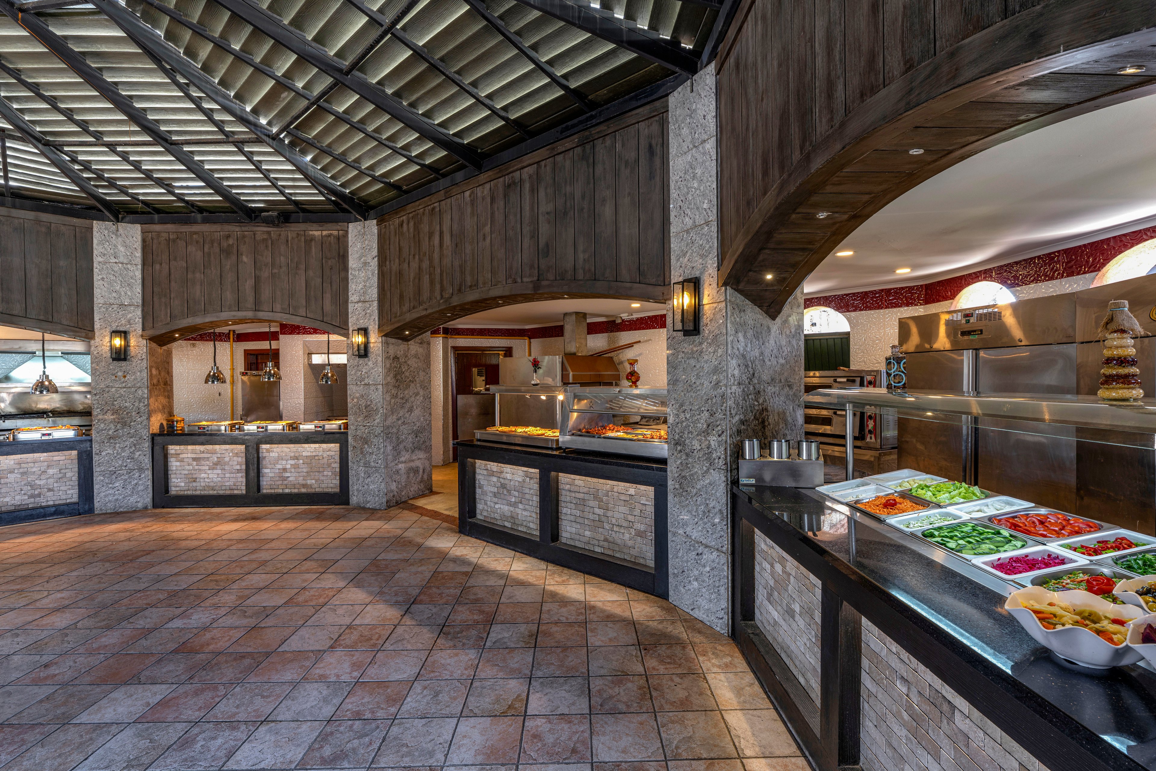 Wide-angle view of a modern restaurant buffet area with multiple food stations. Fresh vegetables and salads are displayed in trays on the right, with colorful ingredients arranged in rows. Warm lighting, tiled floors, and wood-and-stone finishes create a welcoming atmosphere. Stainless steel counters and open serving areas suggest a self-service dining experience.