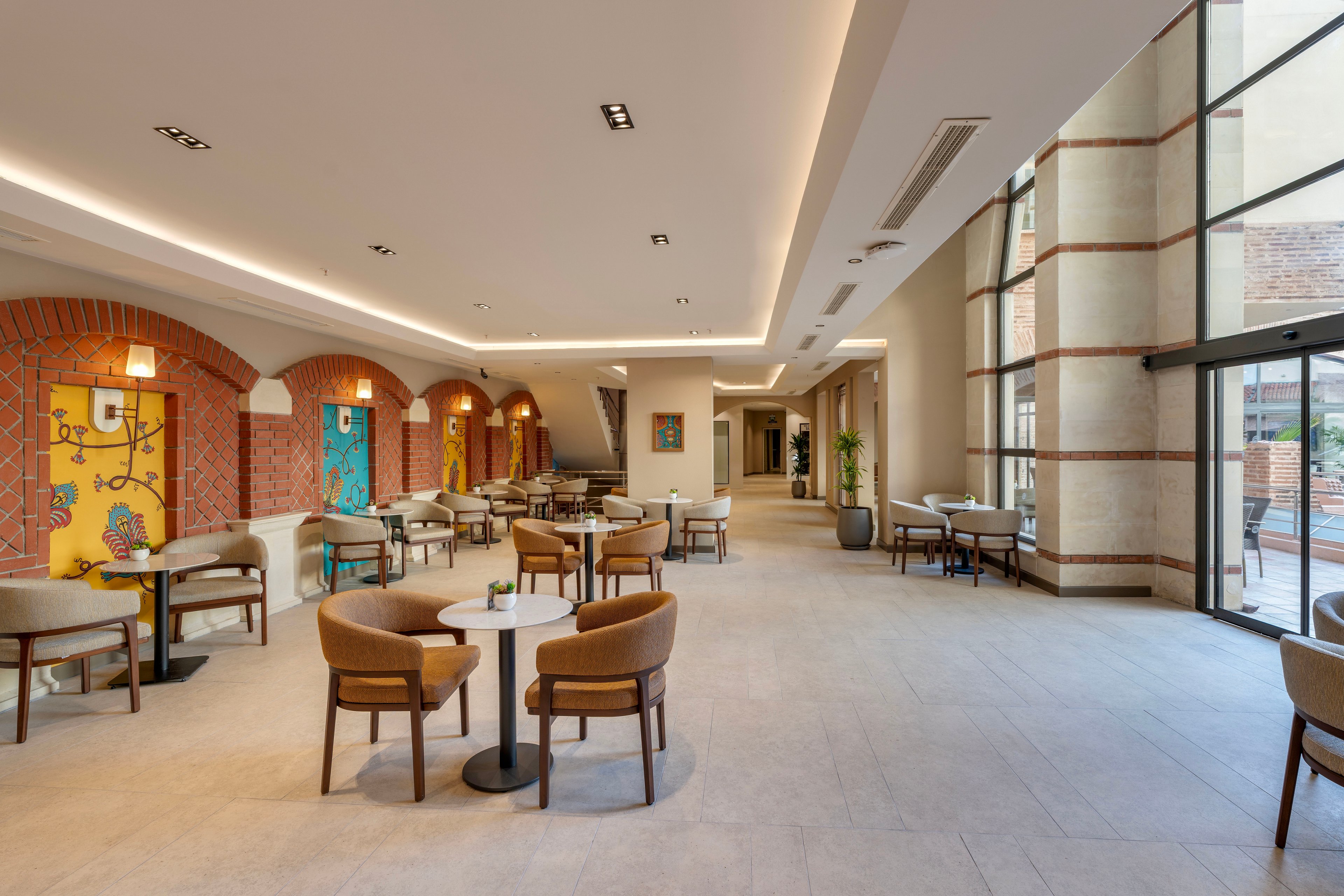 Spacious contemporary lounge area with multiple tables and upholstered chairs arranged for casual seating. The setting features arched brick walls accented by bright, colorful artwork panels and warm pendant lighting. Large floor-to-ceiling windows on the right side let in natural light and offer a view of an outdoor terrace. Neutral flooring and ceiling details create a clean, modern atmosphere suitable for relaxation or informal meetings.