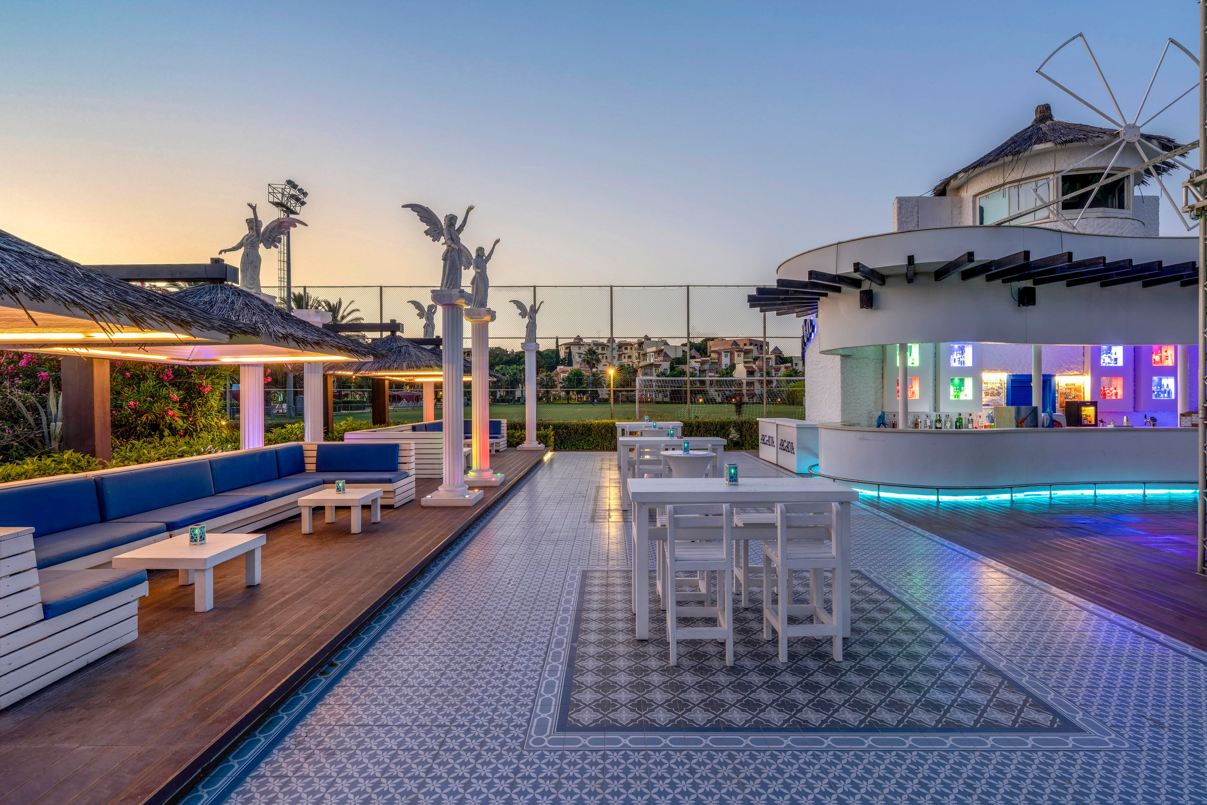 Contemporary open-air bar area with white furniture is shown under a clear evening sky. A curved bar counter with illuminated shelves and colorful backlighting stands to the right. Long blue cushioned benches and low tables line the left side, facing a patterned tile floor and central high tables. Decorative statues and surrounding greenery frame the space, with a distant view of buildings beyond the fence.
