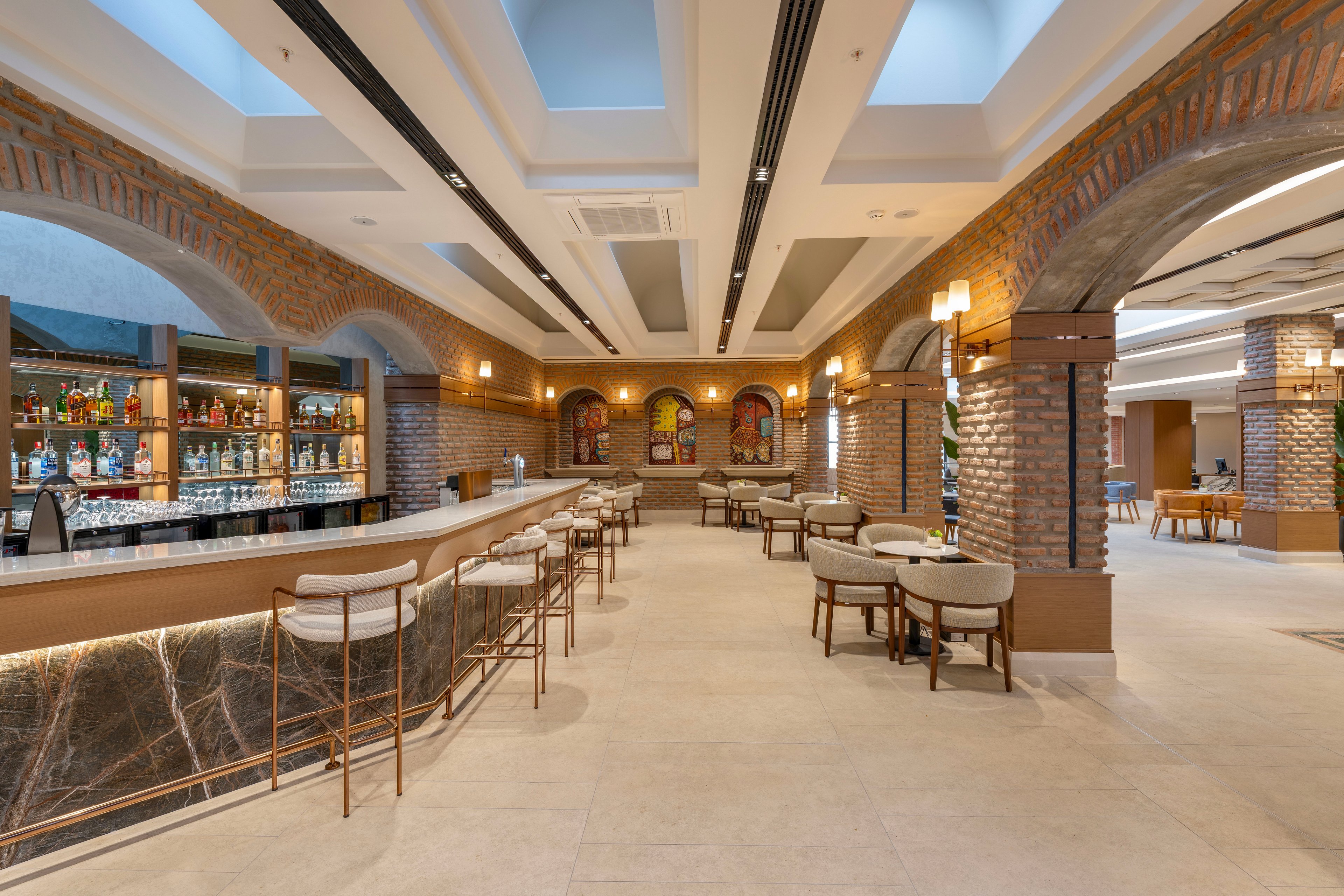 A contemporary bar lounge features a long marble counter lined with sleek metal barstools. Brick arches and columns frame the space, creating a warm, inviting atmosphere with soft ambient lighting. Shelves behind the bar display an assortment of bottles and glassware, while cushioned chairs and small tables provide additional seating. The high ceiling with skylights and recessed lighting enhances the open, airy feel of the interior.