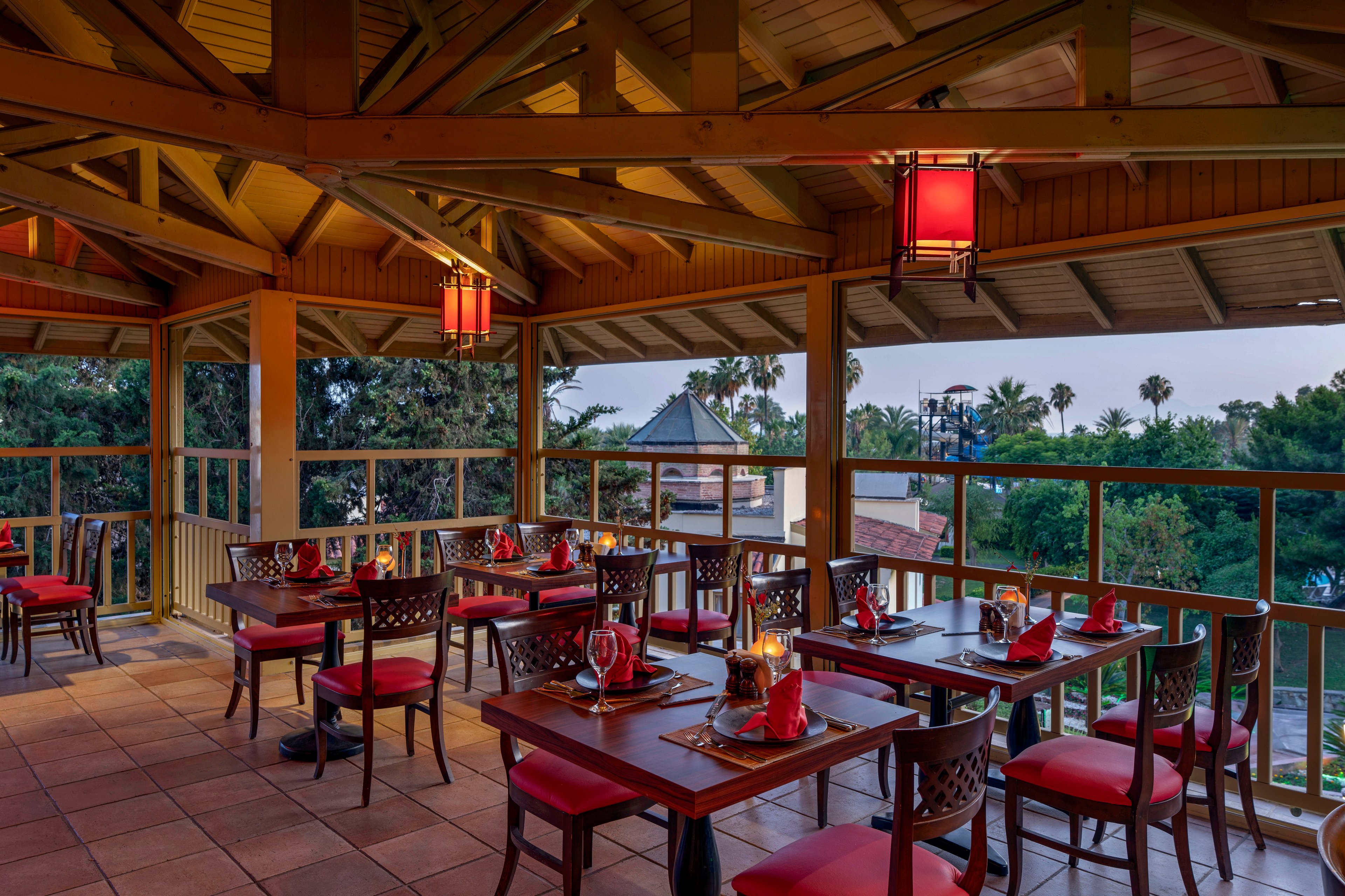 An elevated open-air restaurant terrace features neatly arranged wooden tables with red cushioned chairs and folded red napkins. Warm lantern-style ceiling lights illuminate the space, creating a cozy atmosphere as daylight fades. The terrace overlooks lush green trees and rooftops, framed by wooden railings and exposed beams. Glassware and table settings suggest a prepared dining experience in a relaxed, scenic setting.