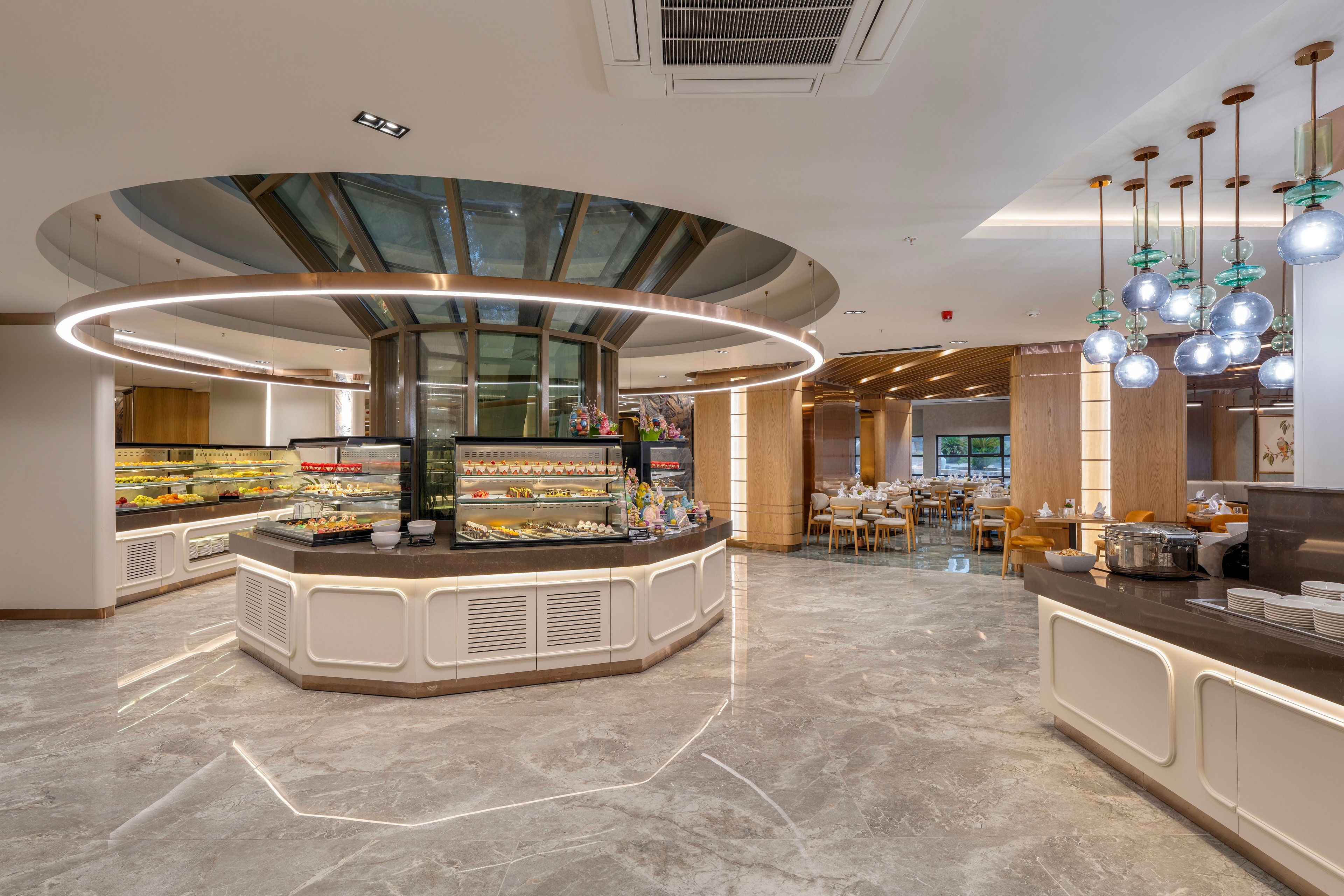Spacious contemporary restaurant interior featuring a central buffet counter with glass display cases of food. The setting includes polished marble flooring, circular ceiling design, and a large ring light above the serving area. Decorative pendant lights with spherical glass shades hang over a side counter, adding warmth to the space. In the background, dining tables and chairs are arranged near large windows, creating an inviting atmosphere for guests.