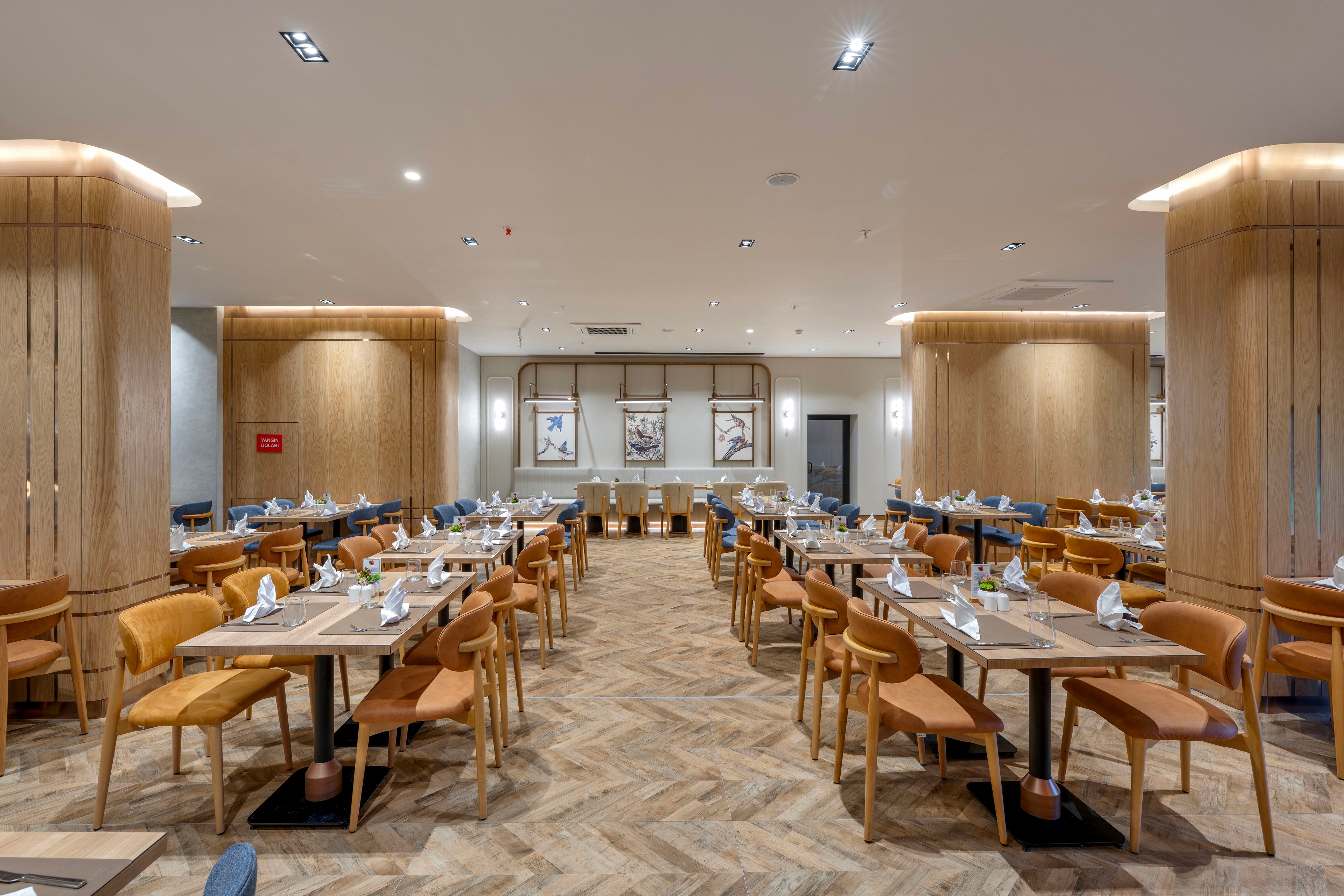 Spacious contemporary restaurant dining room with neatly arranged wooden tables and chairs. The setting features an empty, well-lit space prepared for guests, with napkins and tableware placed on each table. Warm wood tones, leather seating, and herringbone patterned flooring create a clean, modern aesthetic. Large columns, wall art, and recessed ceiling lights add to the sophisticated, welcoming atmosphere.