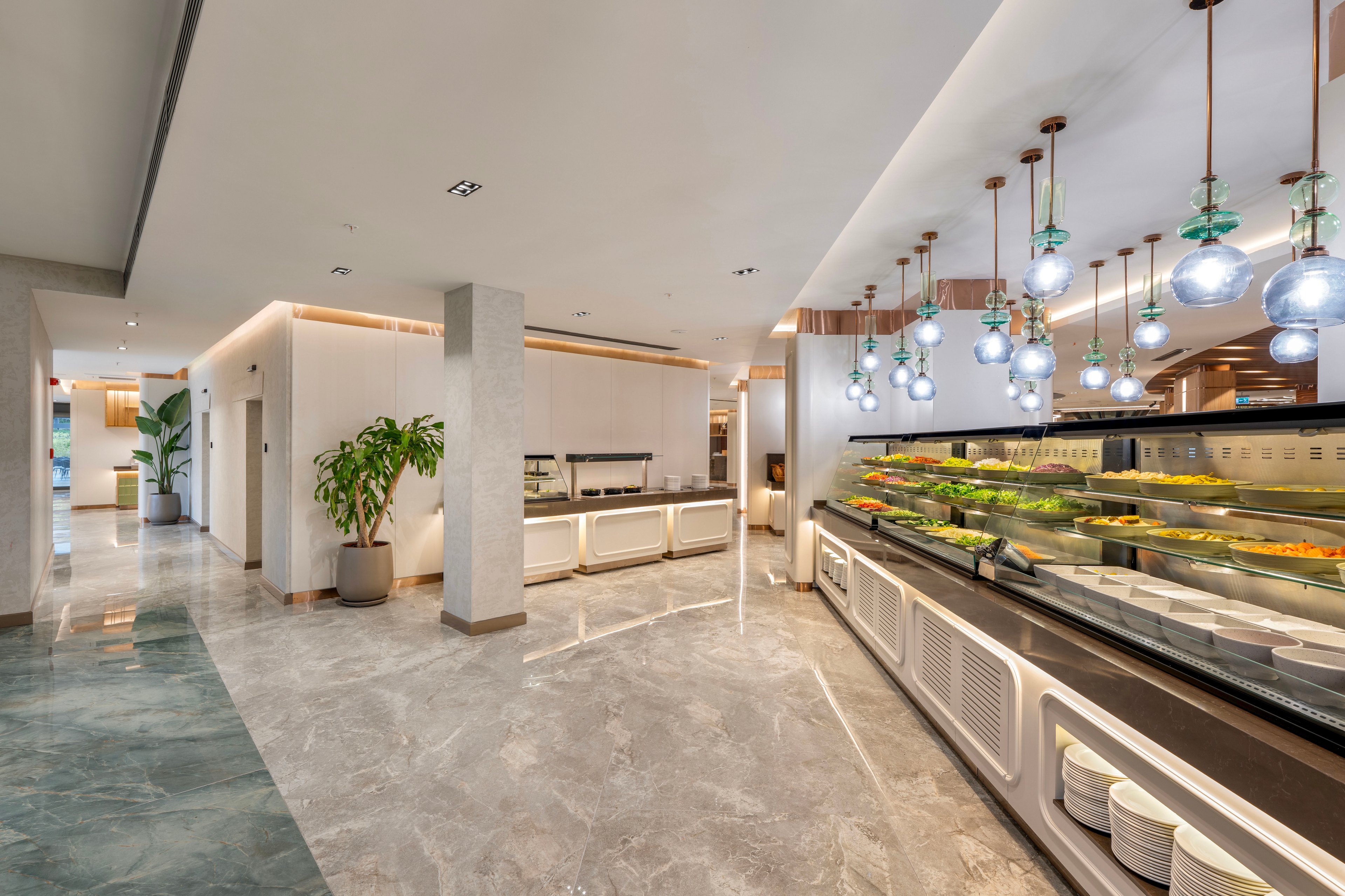 A contemporary buffet area features a long refrigerated salad bar filled with fresh dishes. Pendant lights with spherical glass shades hang above the counter, creating a bright and inviting atmosphere. The space has polished stone flooring, light wood paneling, and neatly stacked white plates beneath the counter. A large potted plant and clean, minimal cabinetry enhance the modern interior design of the restaurant or hotel dining area.
