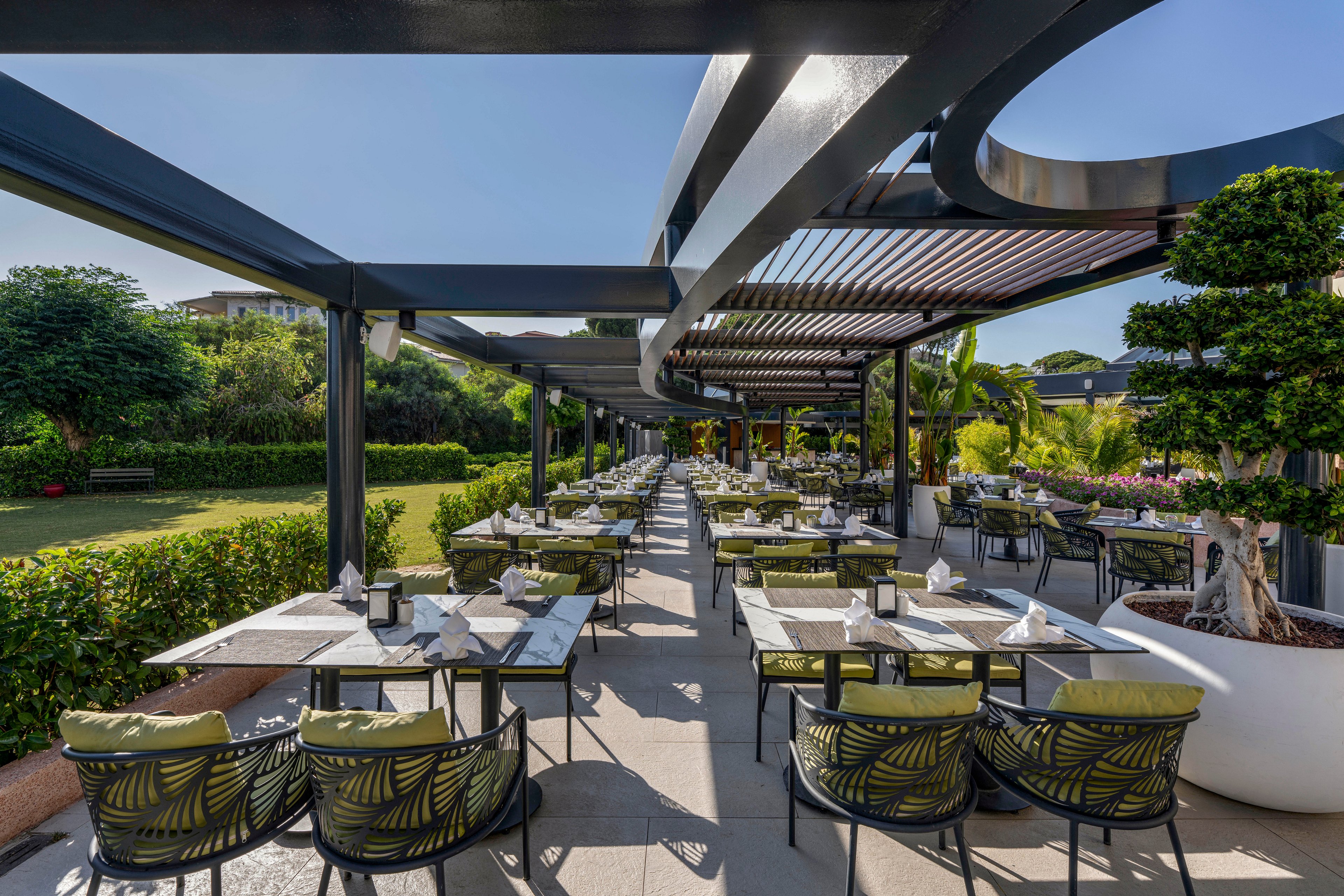 An elegant outdoor restaurant terrace features long rows of tables and chairs arranged beneath modern pergola structures. The setting overlooks a lush landscaped garden with manicured hedges, trees, and flowering plants under a clear blue sky. Tables are neatly set with napkins and tableware, suggesting readiness for guests in a relaxed al fresco dining atmosphere. Contemporary furniture with patterned cushions and large potted plants adds a stylish, resort-like feel to the space.