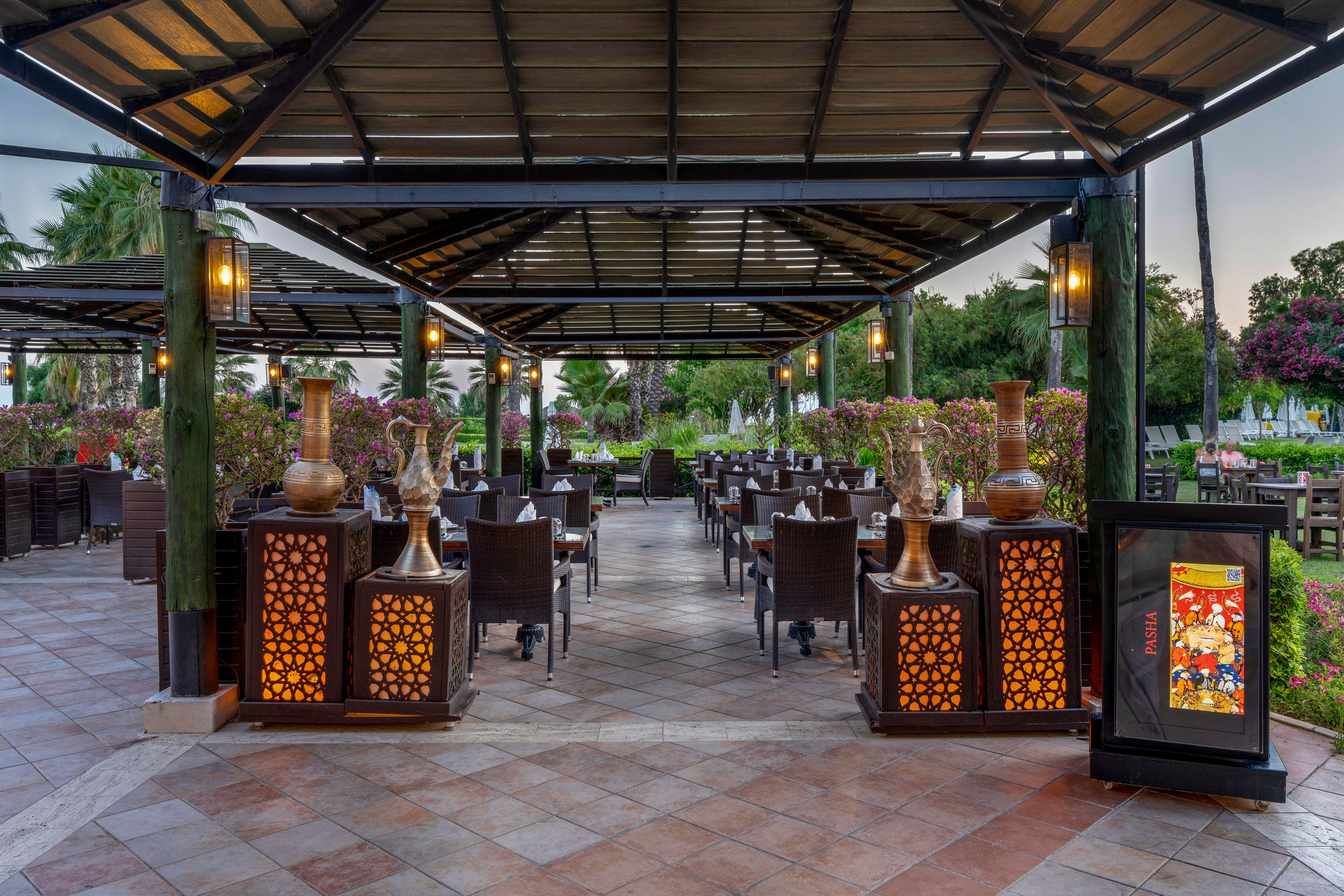 An open-air restaurant terrace features rows of neatly arranged tables and dark wicker chairs under a large metal roof structure. Warm lantern-style lights and ornate illuminated panels create a welcoming ambiance along the central aisle. Decorative vases and plants accent the dining area, while lush greenery and flowering trees surround the space. A standing display board with colorful artwork is positioned near the entrance, inviting guests into the setting.