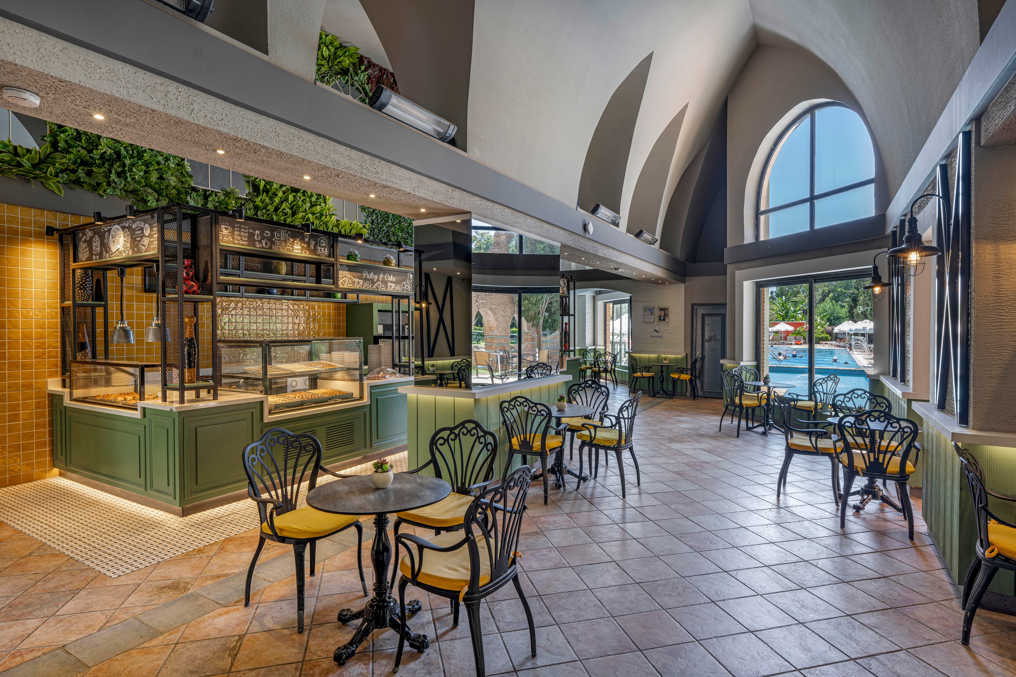 Spacious café interior features small round tables with decorative metal chairs and yellow cushions. A stylish service counter with glass display and green cabinetry stands to the left, framed by warm tile and greenery accents. Large arched windows and glass doors let in abundant natural light, revealing an outdoor pool area beyond. The tiled floor and high vaulted ceiling create an airy, contemporary atmosphere suitable for casual dining or coffee breaks.