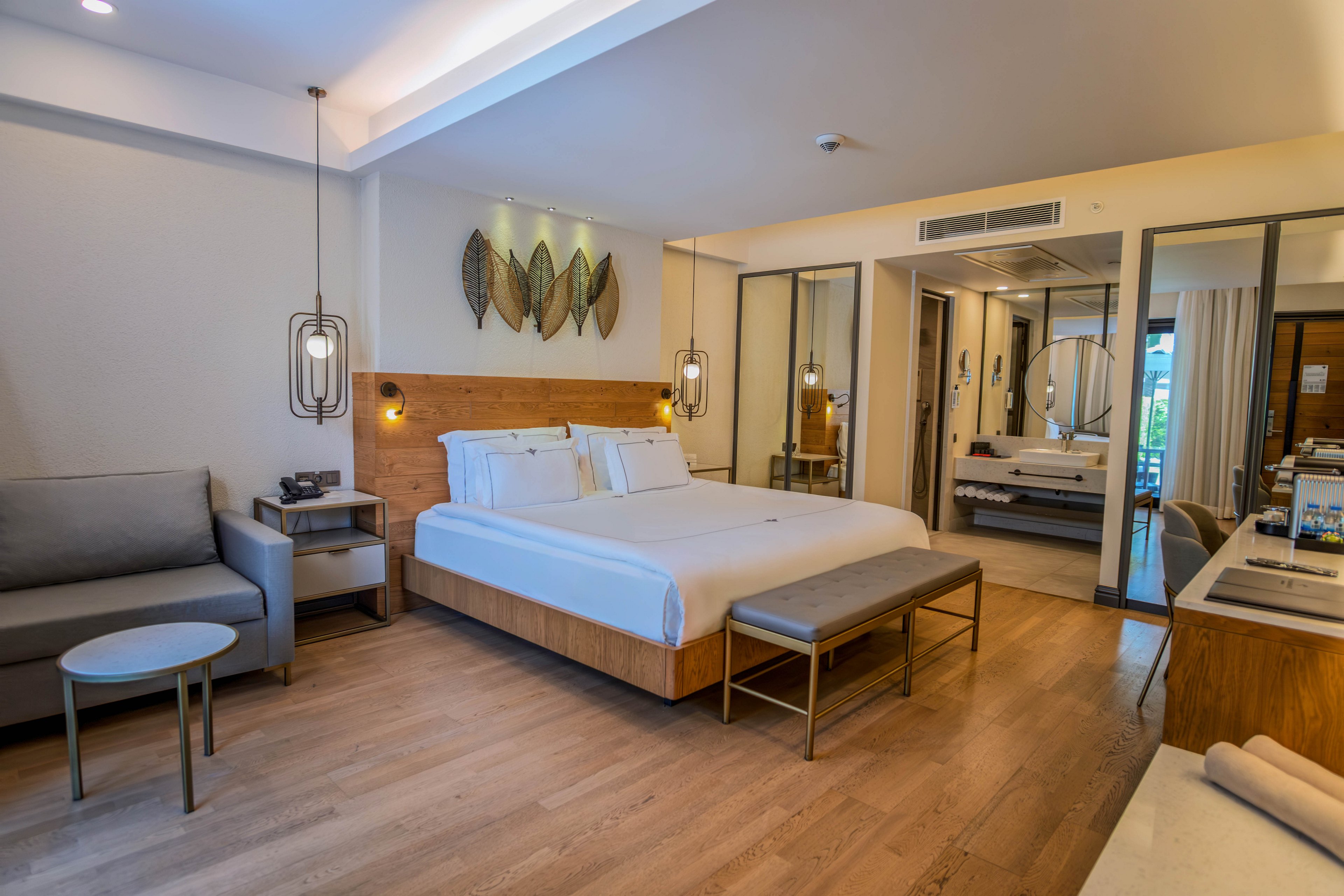 A spacious modern hotel room features a large bed with crisp white linens and a wooden headboard. The setting includes a gray sofa, small round side table, and a bench at the foot of the bed. Warm pendant lights and decorative wall art create a cozy, contemporary atmosphere. Large mirrors and a desk area enhance the sense of space and functionality in the room.