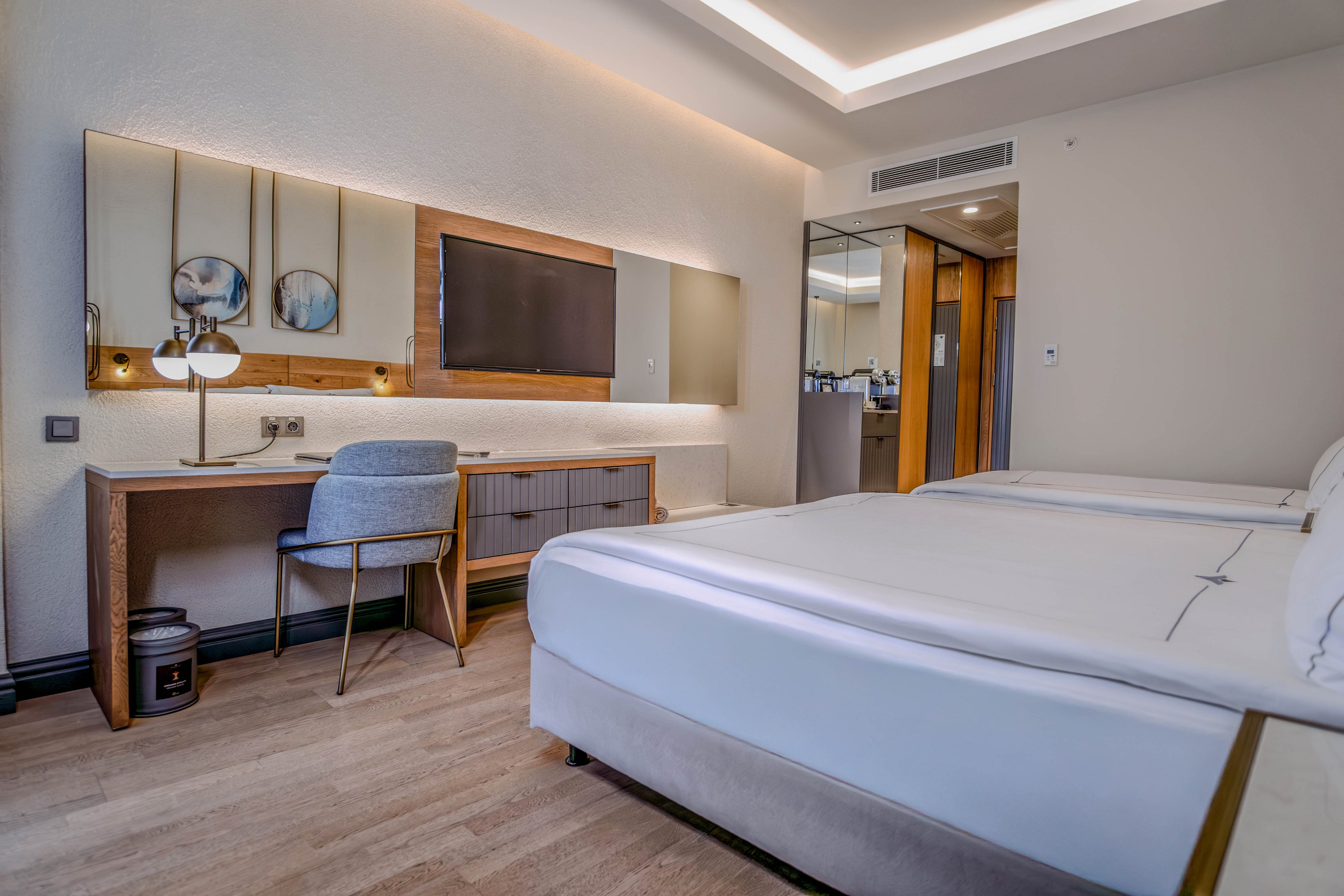 Contemporary hotel room interior featuring two neatly made twin beds with white linens. A wooden desk with a cushioned chair and table lamp sits opposite the beds beneath a large wall-mounted television and mirror. Warm recessed ceiling lighting and light wood flooring create a clean, inviting atmosphere. A doorway leads to the entrance and bathroom area, emphasizing the functional layout of the space.