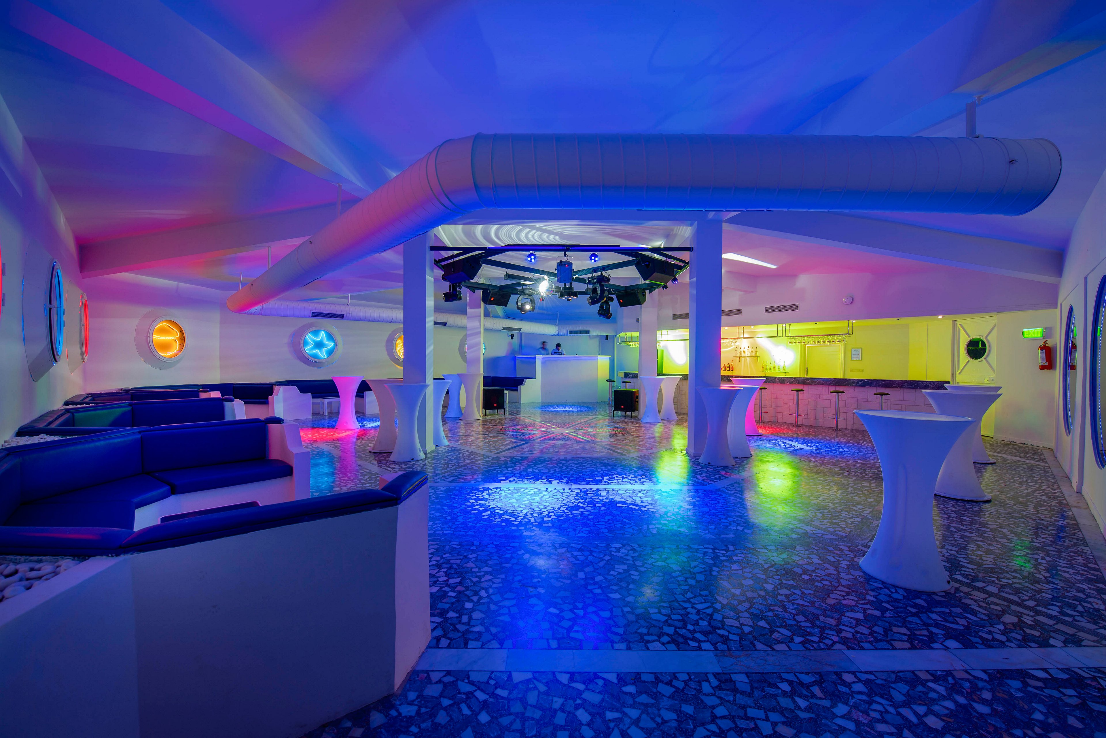 A spacious modern lounge interior is illuminated with vibrant blue, purple, and pink club lighting. Curved sofas line the walls while tall cocktail tables stand in the center of the room. The terrazzo-style floor reflects the colorful lights, adding to the futuristic atmosphere. The space appears ready for an event or party, with a bar area and ceiling-mounted lighting equipment visible in the background.