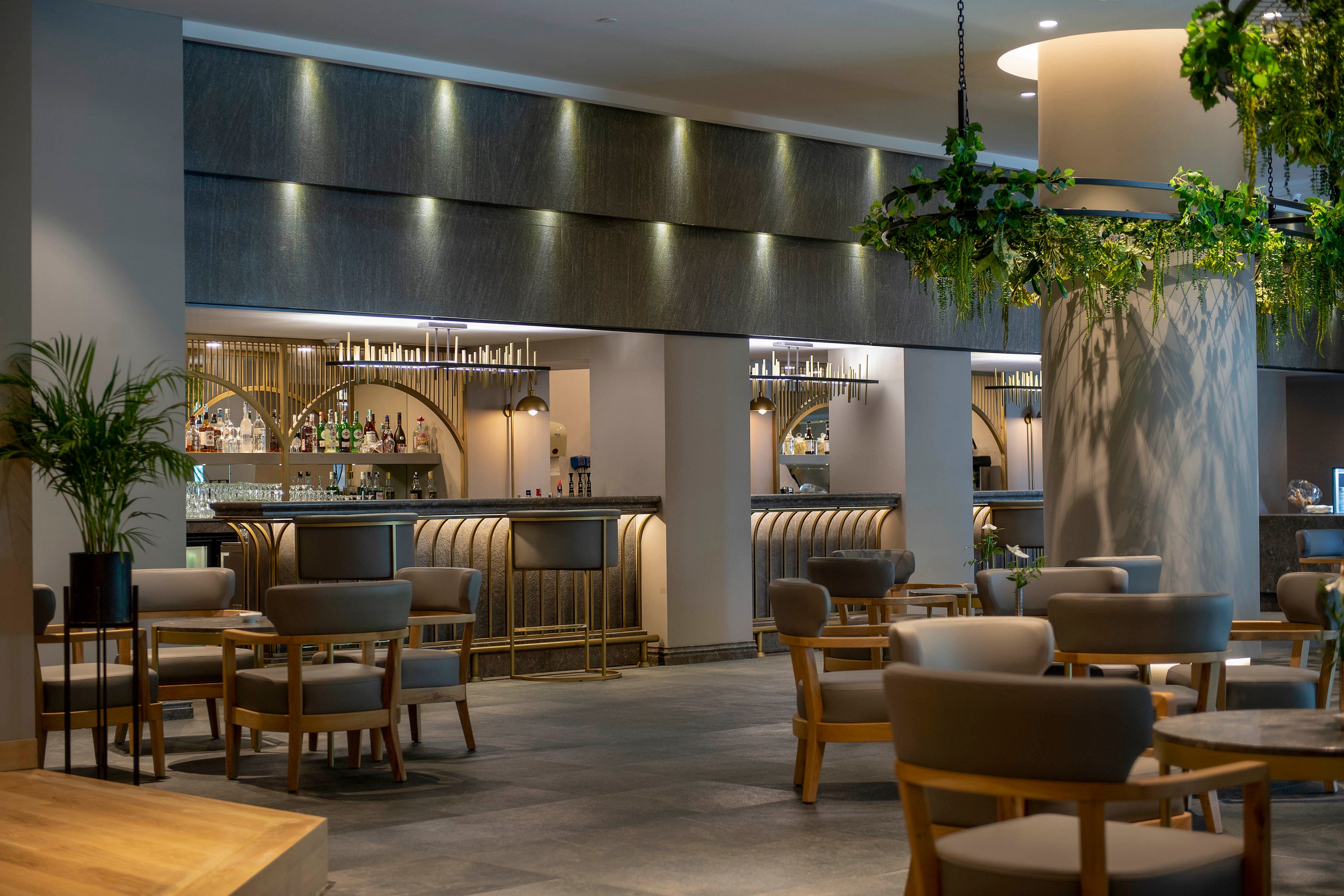A stylish hotel bar lounge features a long counter with backlit shelves of bottles and glassware. Contemporary armchairs and small tables are arranged across the open floor, creating an inviting social space. Soft, indirect lighting highlights the bar area and textured wall panels. Green plants and hanging foliage add a touch of nature to the otherwise minimalist, neutral-toned interior.
