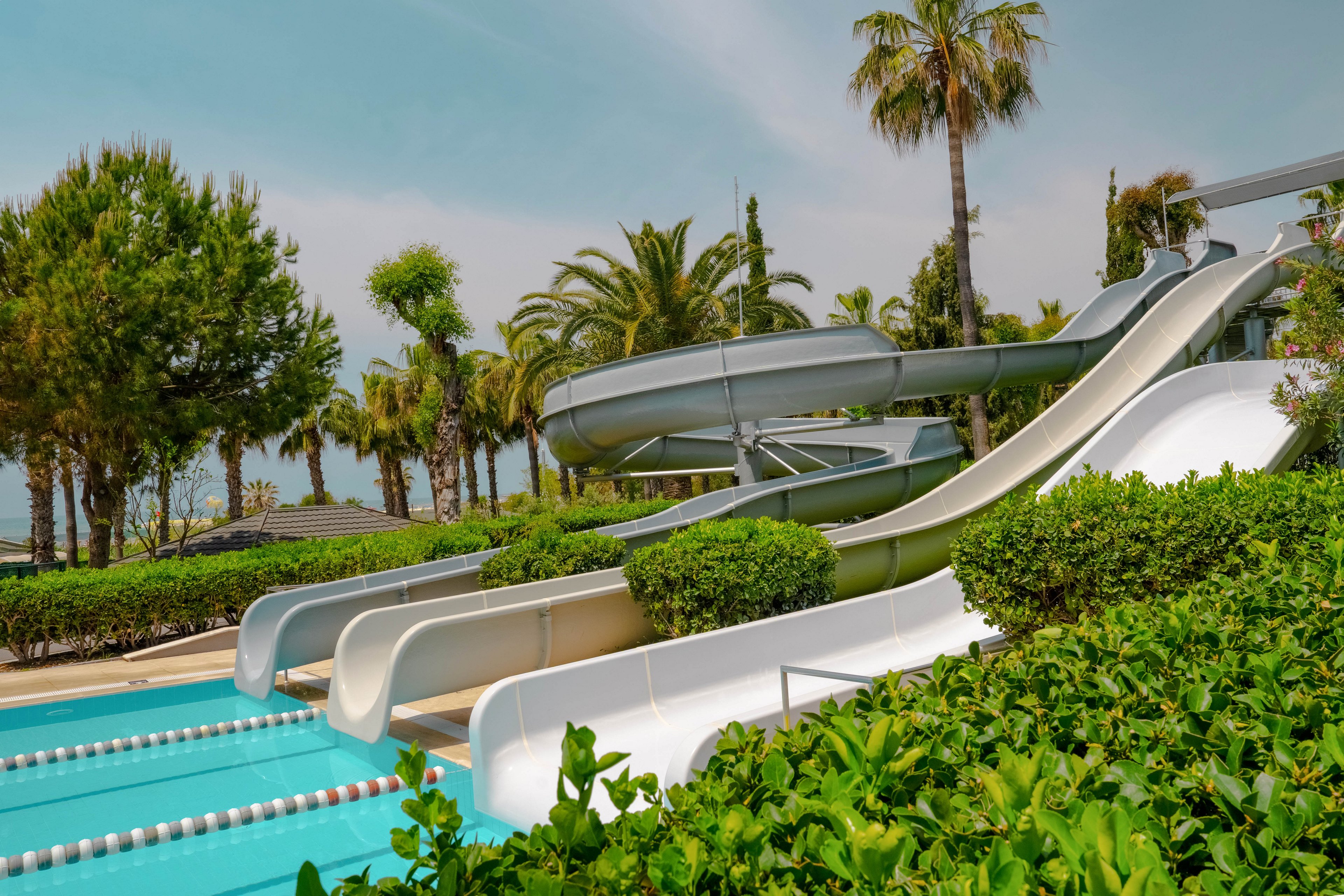 An outdoor water park features several white and gray water slides leading into a bright blue swimming pool. The scene is surrounded by lush green hedges and tall palm trees, suggesting a warm, tropical climate. The sky is clear with a light haze, enhancing the relaxed vacation atmosphere. No people are visible, emphasizing the clean lines and structure of the slides and pool area.